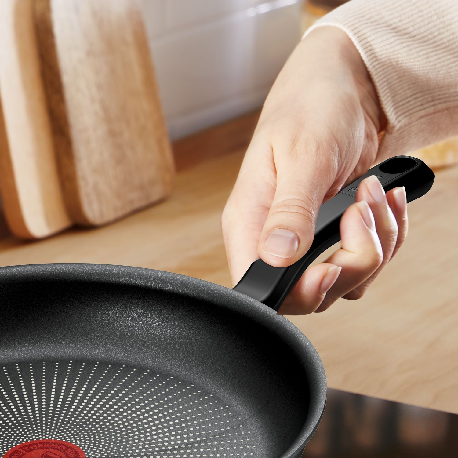 Hand holding a black frying pan with a red dot, wooden cutting boards in background.