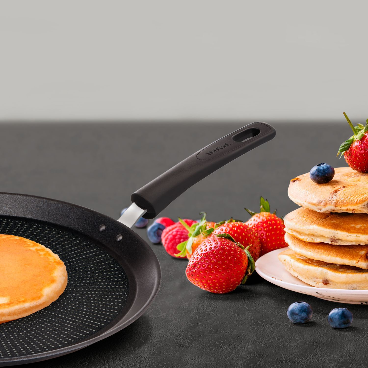 Pancakes in a pan with berries and a stack of pancakes on a plate.