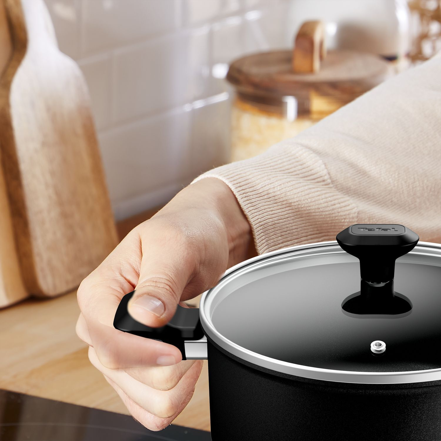 A person's hand holding the handle of a black pot with a glass lid.