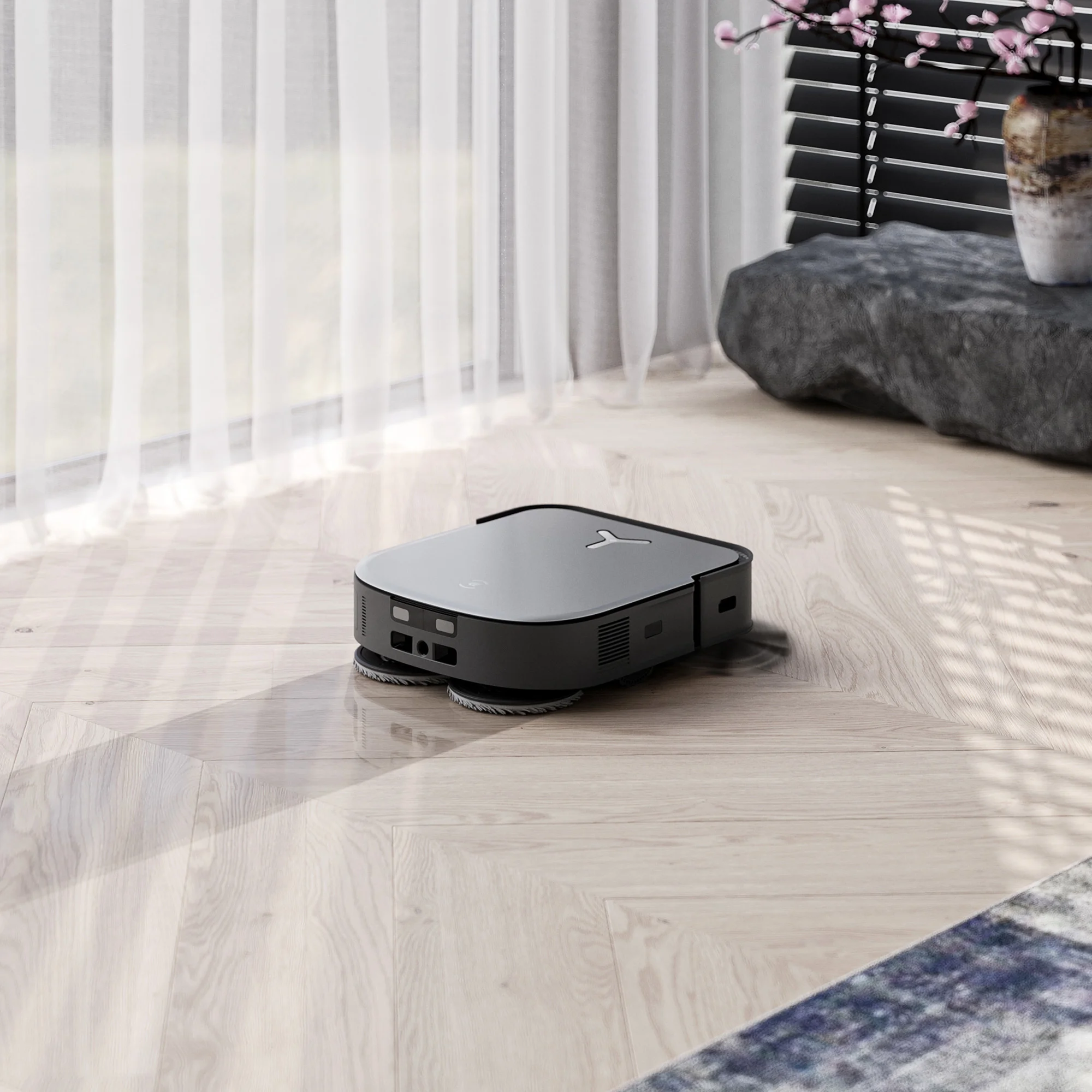 Un aspirapolvere robot grigio su un pavimento in legno. Lo sfondo mostra una finestra, tende e un vaso.