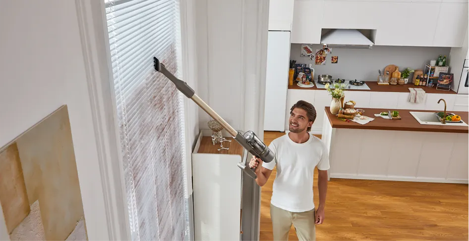 Un homme passe l'aspirateur sur des stores avec un aspirateur sans fil dans une cuisine moderne.