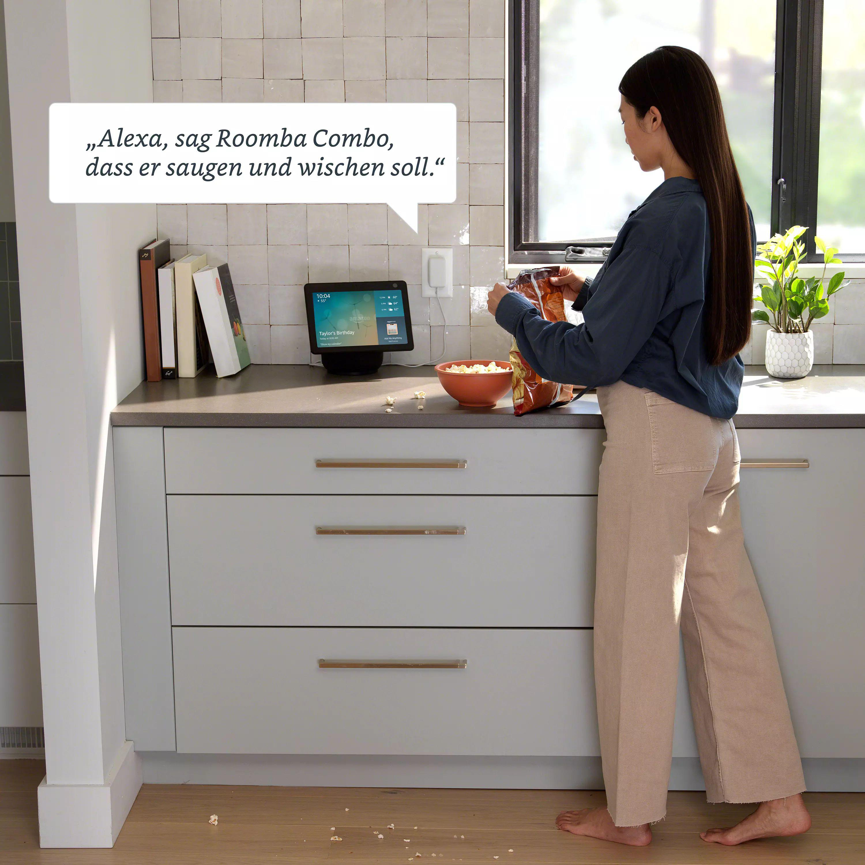 Eine Frau in einer Küche mit einem Smart Display; Text sagt, Alexa soll dem Roomba sagen, er soll putzen.
