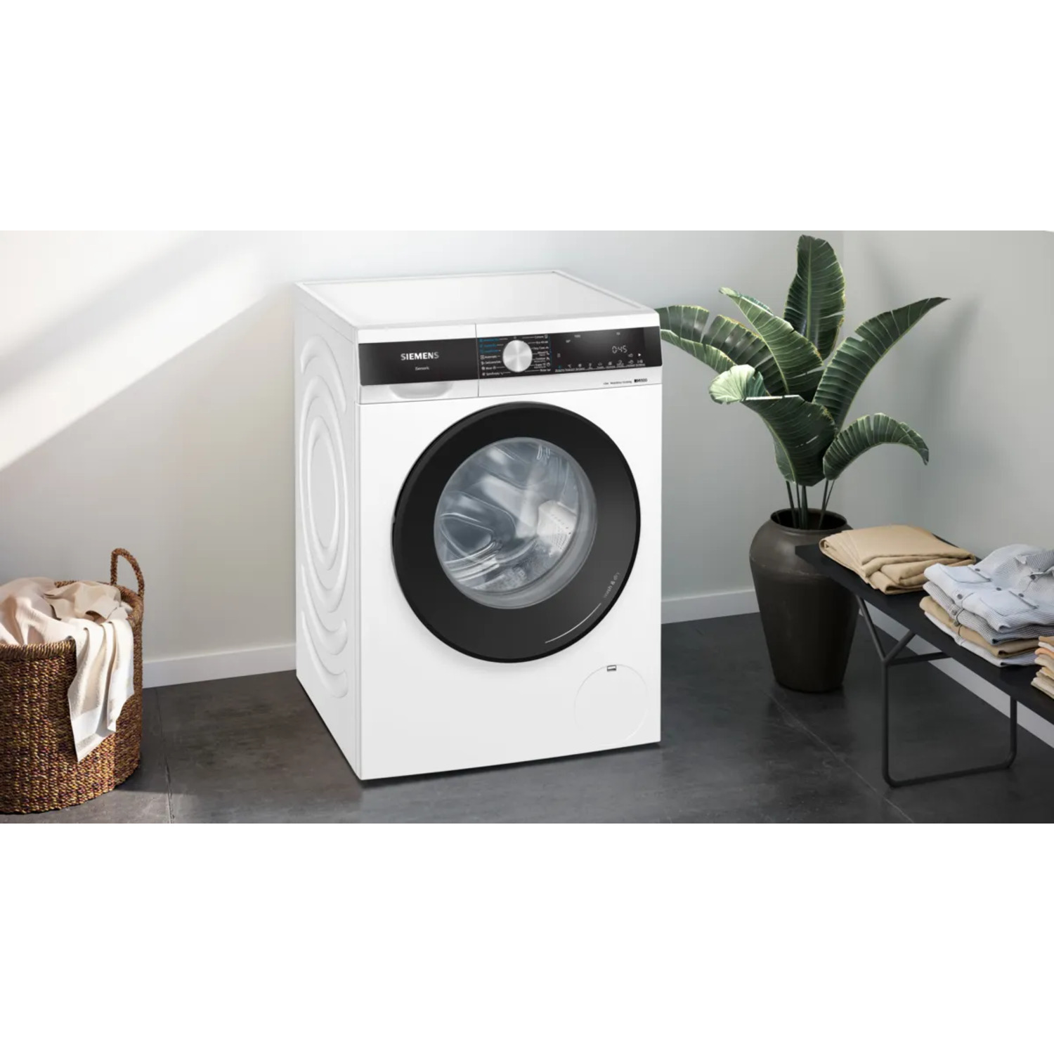 White washing machine with black door, near a plant and basket.