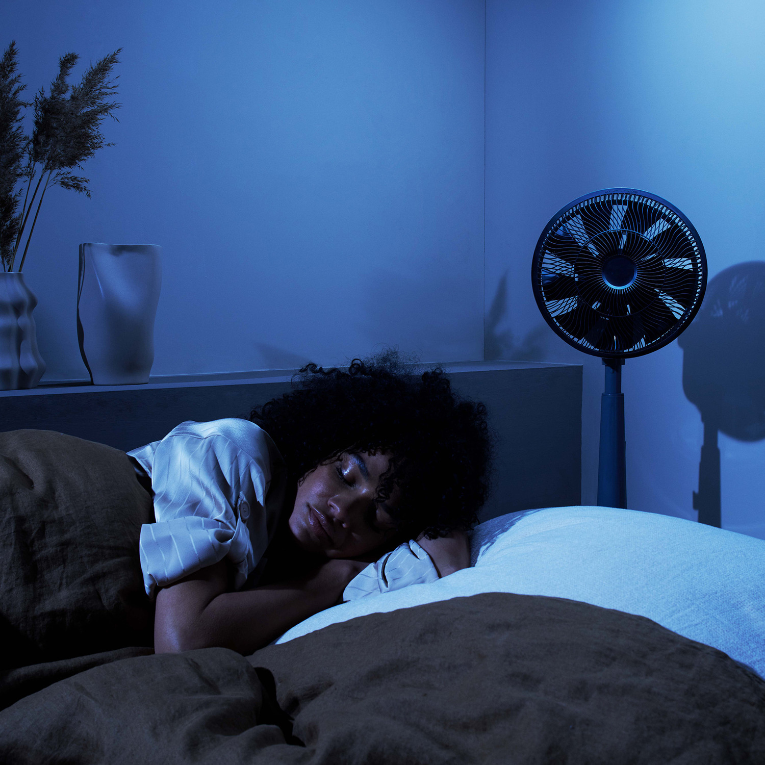 Een vrouw die slaapt in een bed met een ventilator, een vaas met bloemen en een blauwe muur.