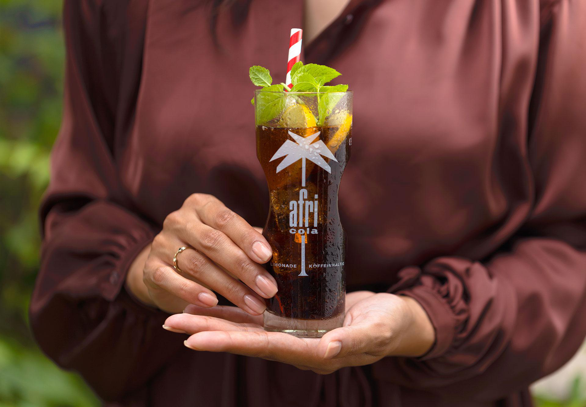 Person hält ein Glas Afri-Cola, garniert mit Minze und Strohhalm. Braunes Kleid, grüner Hintergrund.