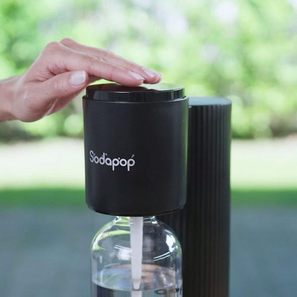 Hand auf schwarzem SodaPop-Spender. Klare Glasflasche. Hintergrund verschwommen grün.