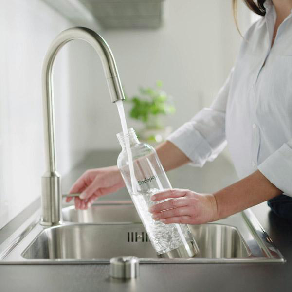 Eine Frau füllt eine Flasche mit Wasser aus einem Wasserhahn in einem Edelstahlbecken. Das Wasser ist klar.