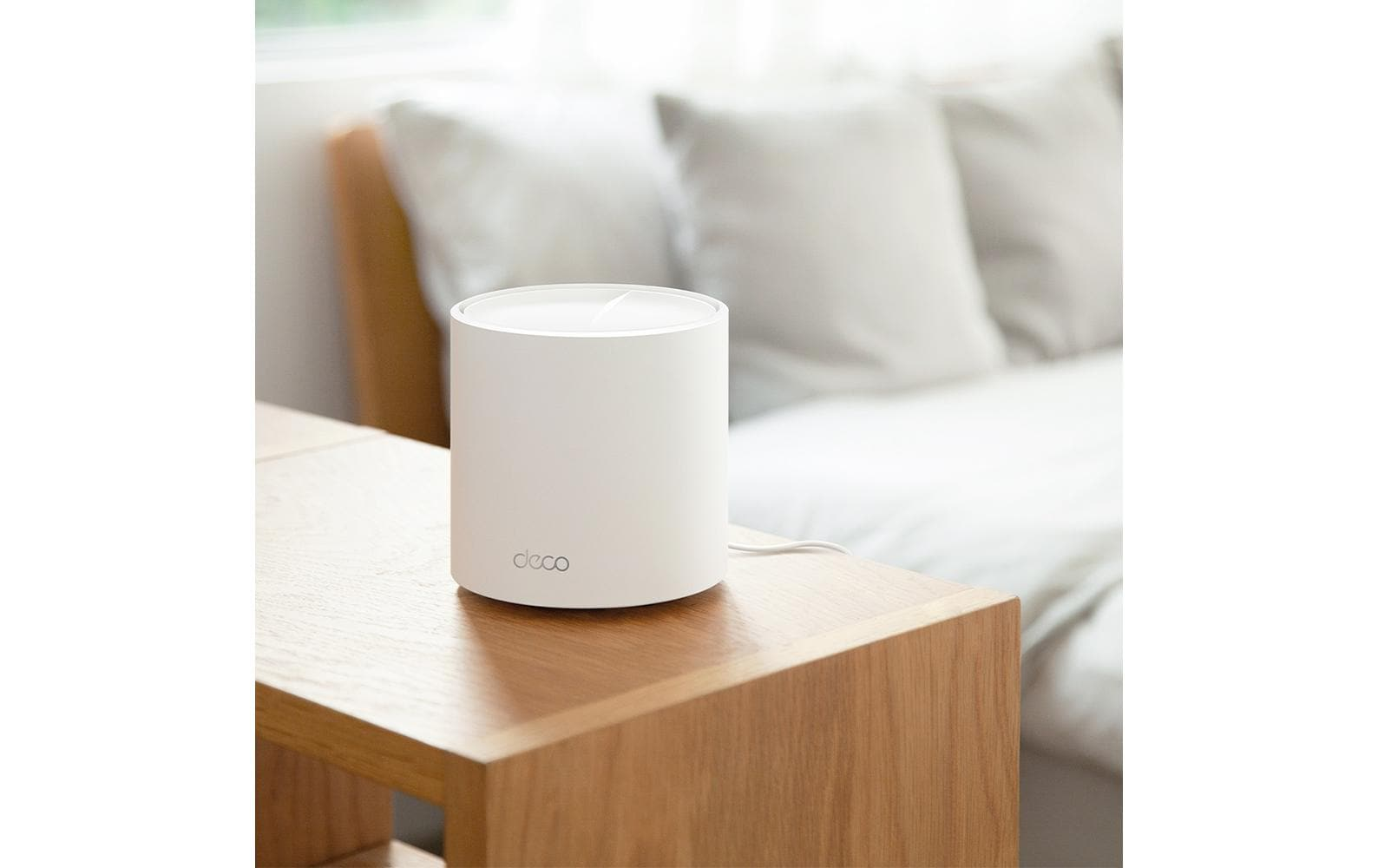 Un modem cylindrique blanc est posé sur une table en bois. Derrière, on aperçoit un lit avec des oreillers blancs.