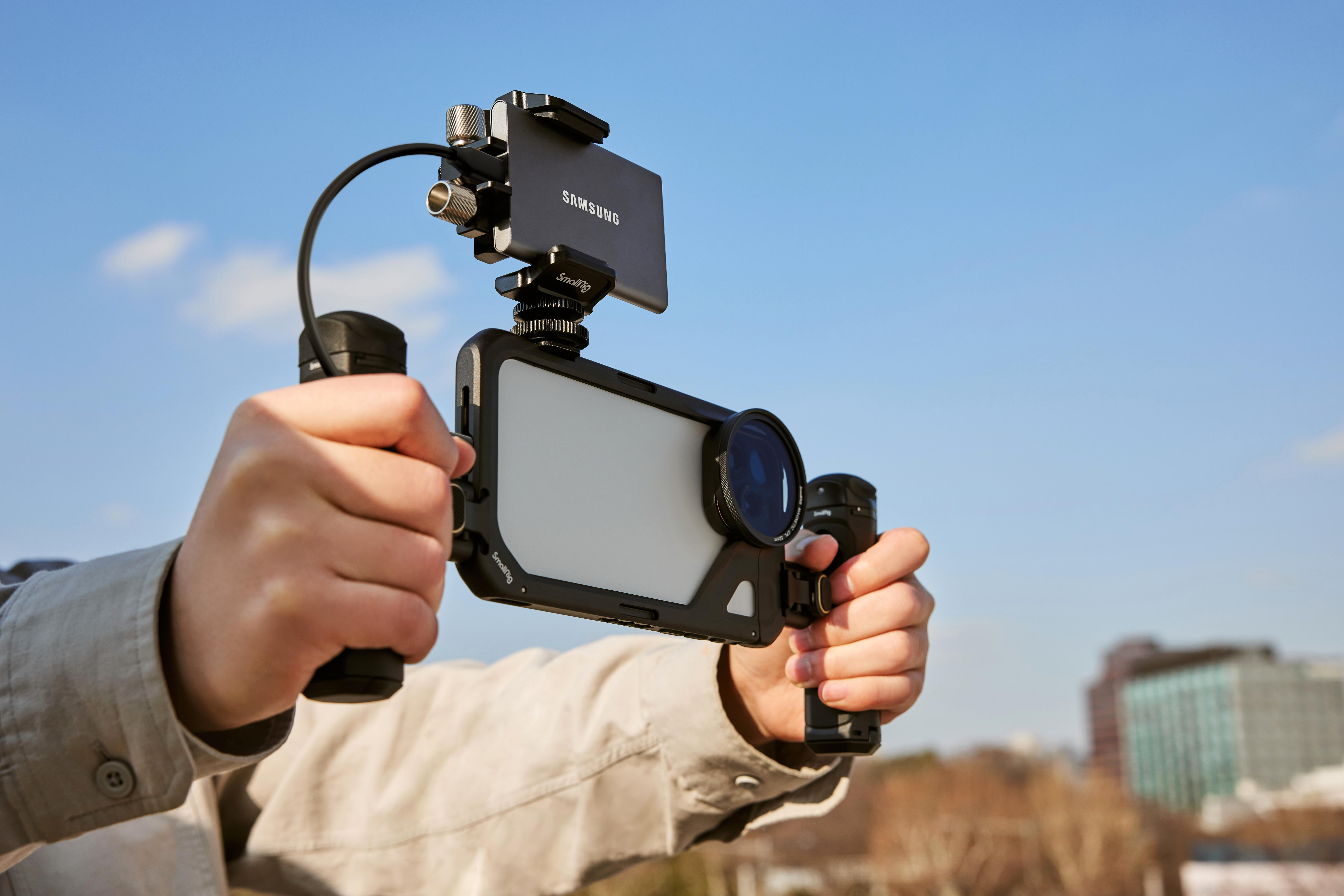 Hände halten Smartphone mit Rig für Videoaufnahmen, blauer Himmel im Hintergrund.