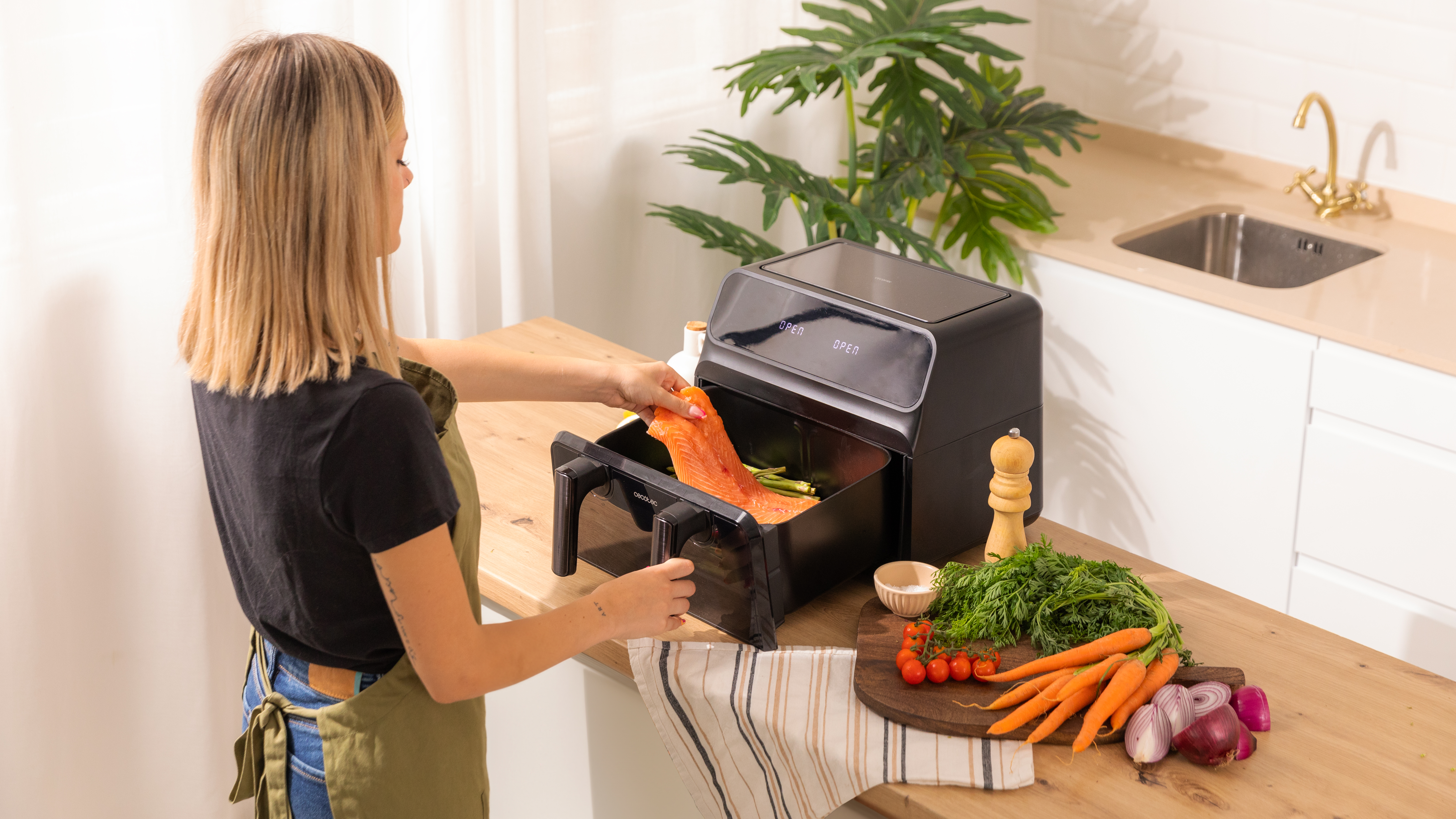 Una mujer pone salmón y espárragos en una freidora de aire junto a una mesa de madera con verduras.