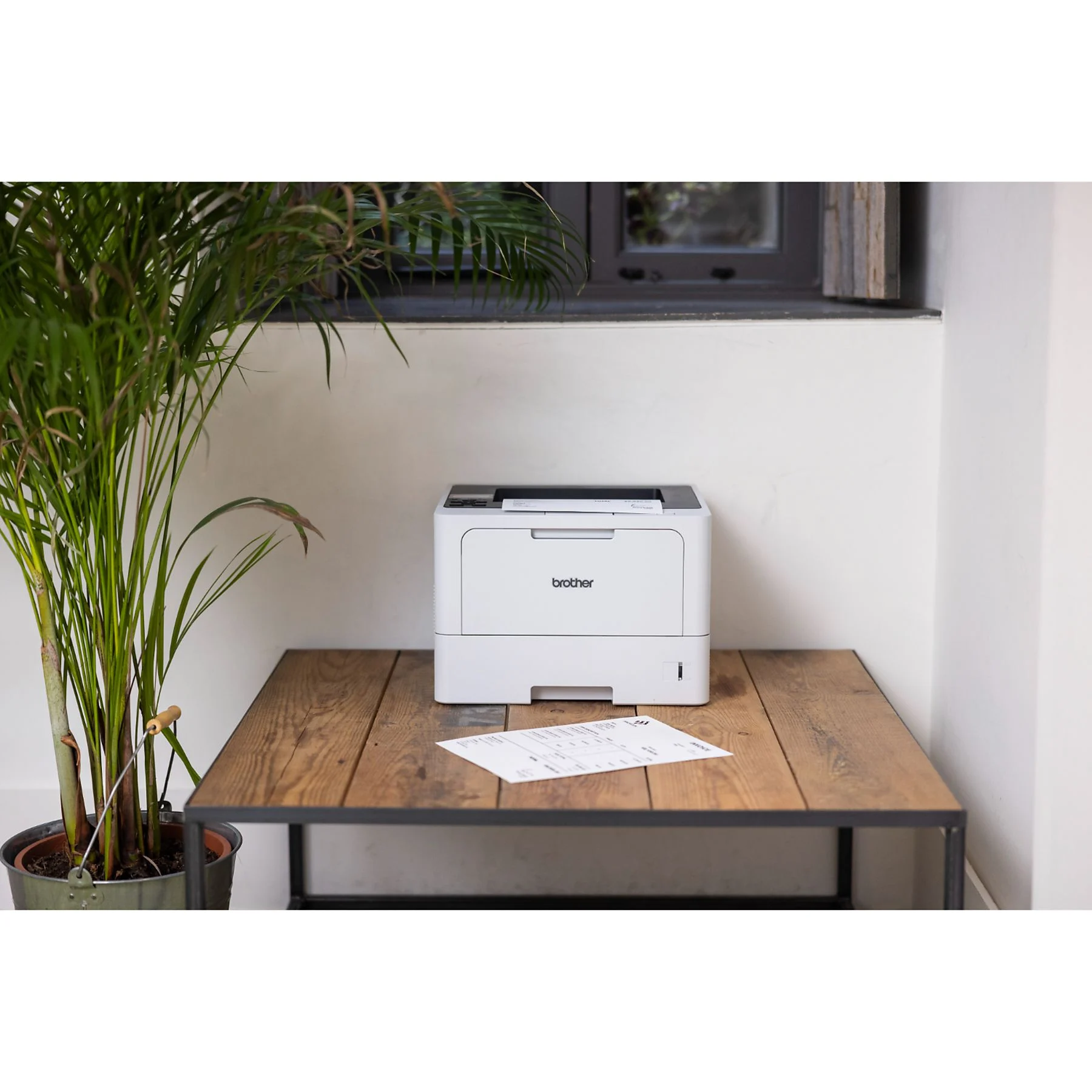 Una impresora Brother blanca sobre una mesa de madera. Una planta a la izquierda. Un documento en la mesa.