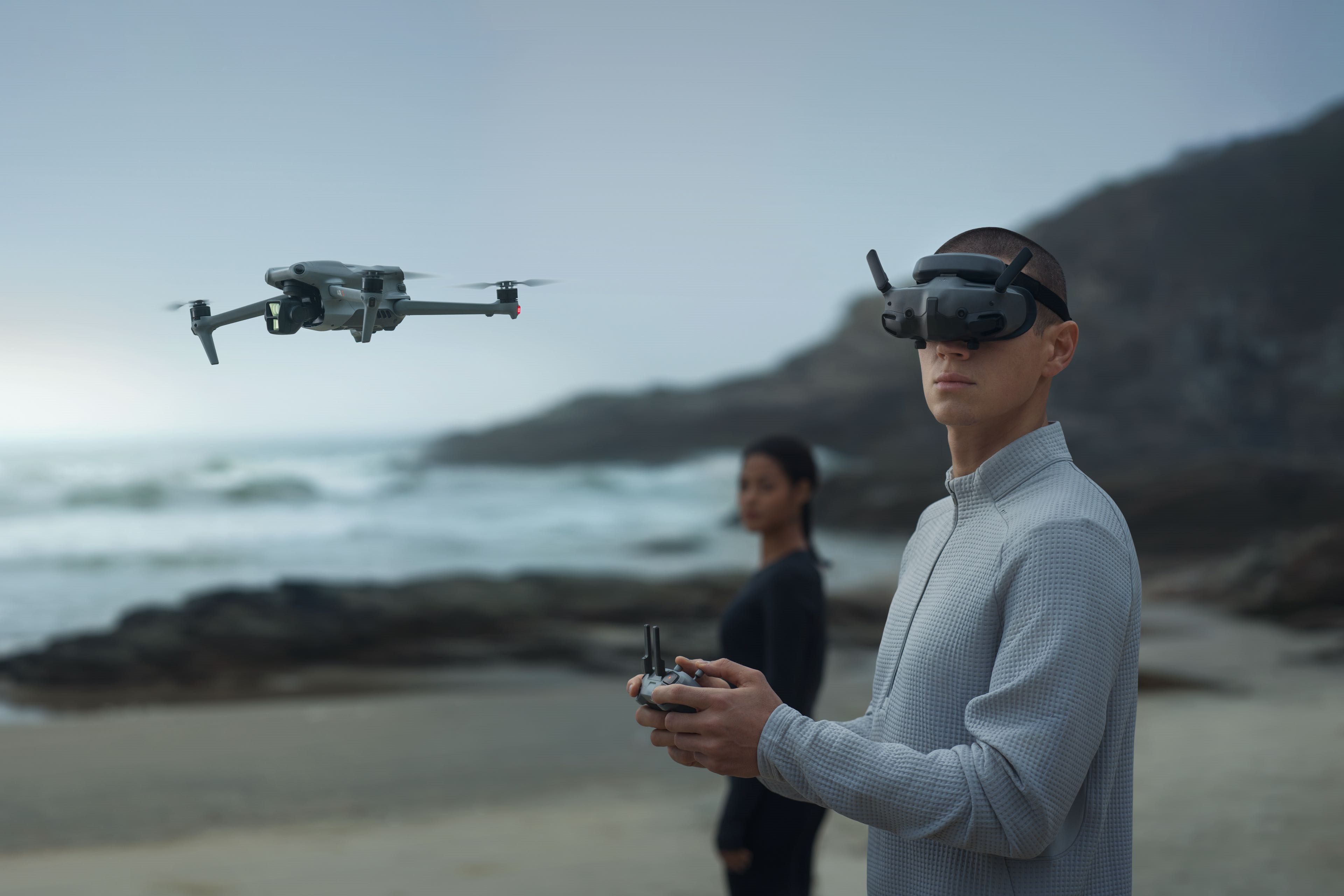 Mężczyzna w goglach VR i z kontrolerem obsługuje drona nad plażą. Za nim stoi druga osoba.