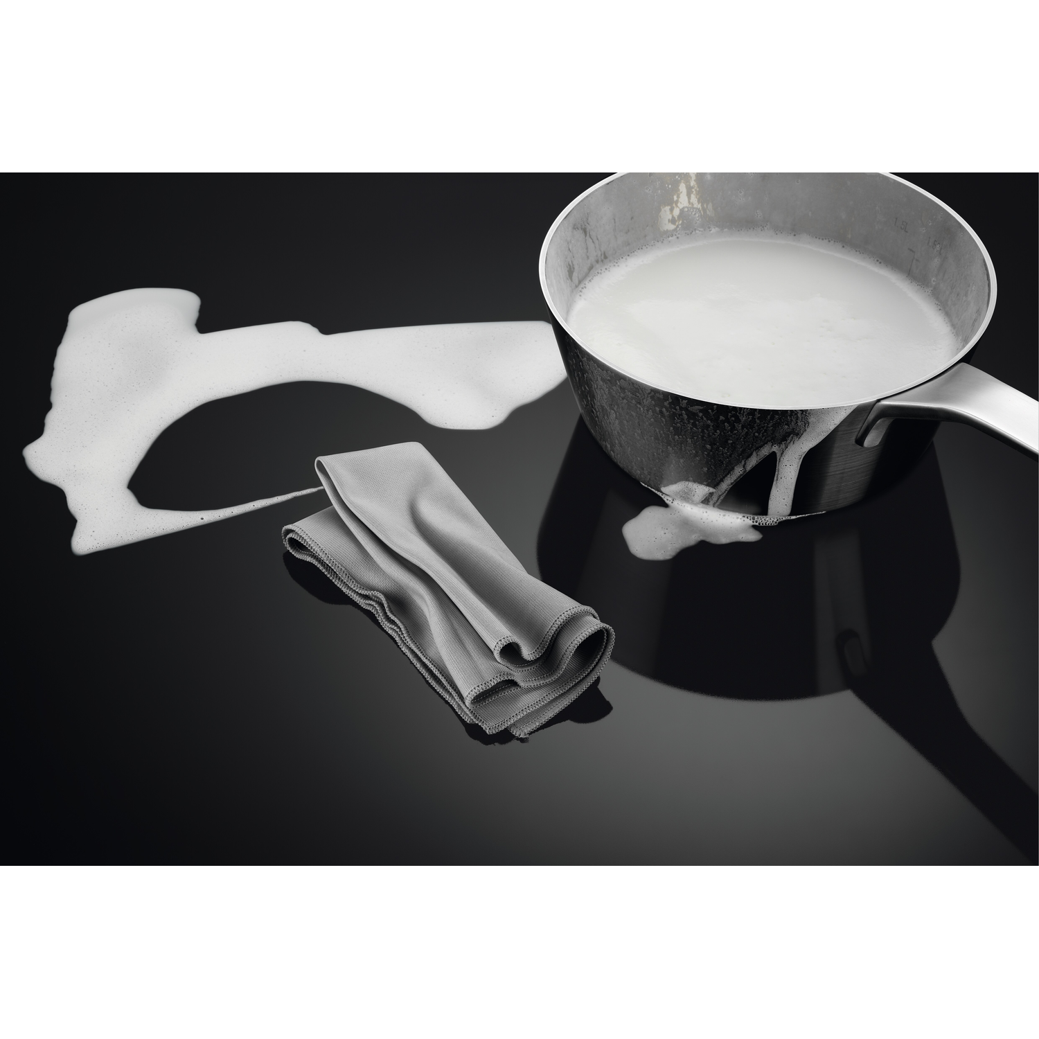 Een roestvrijstalen pan met overkokende melk, op een zwart oppervlak. Een grijze doek ligt naast de pan.