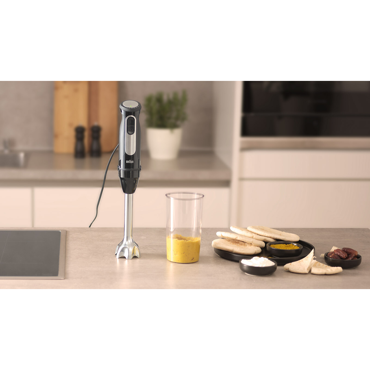 Staafmixer in een keuken met een glas gele vloeistof, pitabrood en andere ingrediënten.