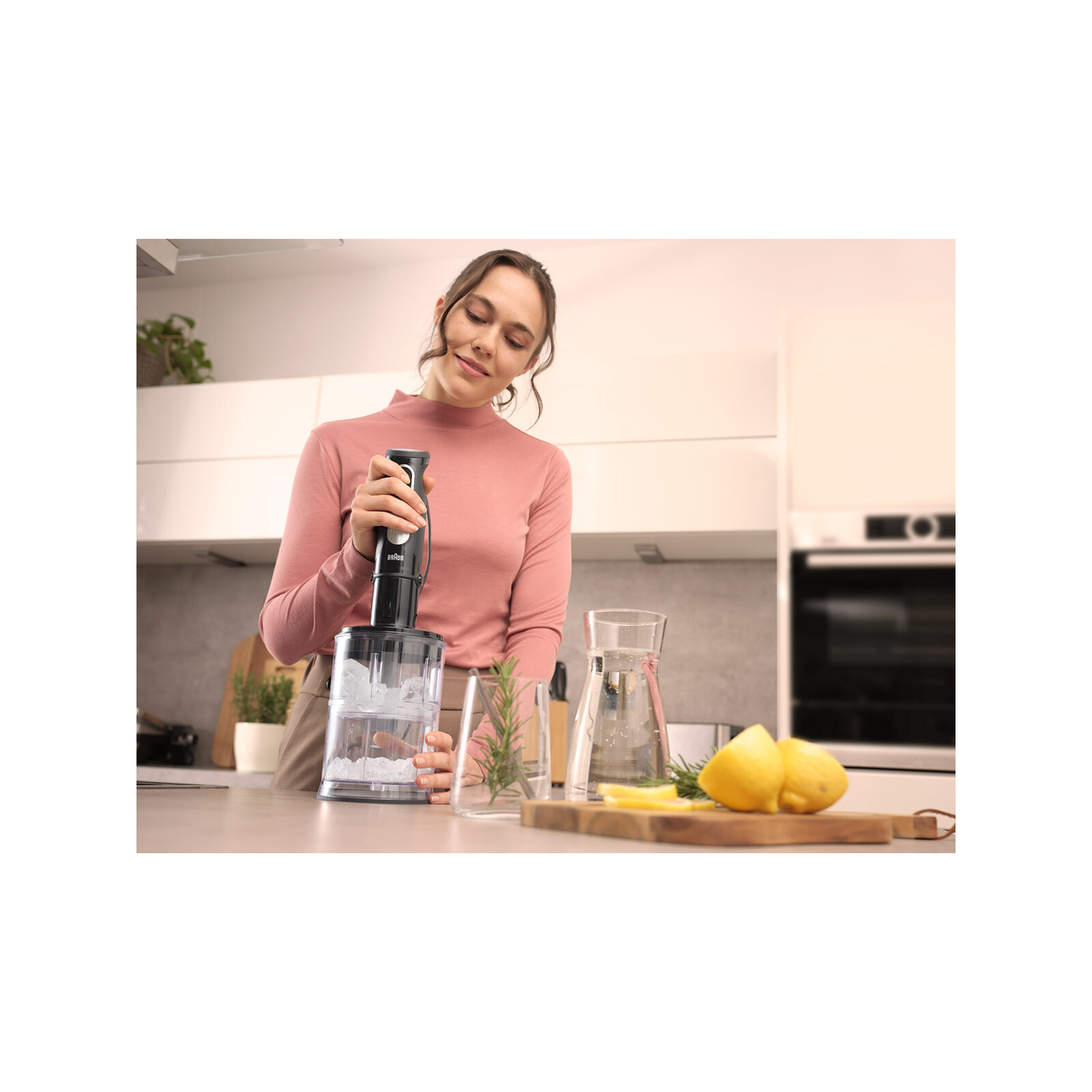 Een vrouw gebruikt een staafmixer in een glas water met ijs, citroenen en rozemarijn in de buurt.