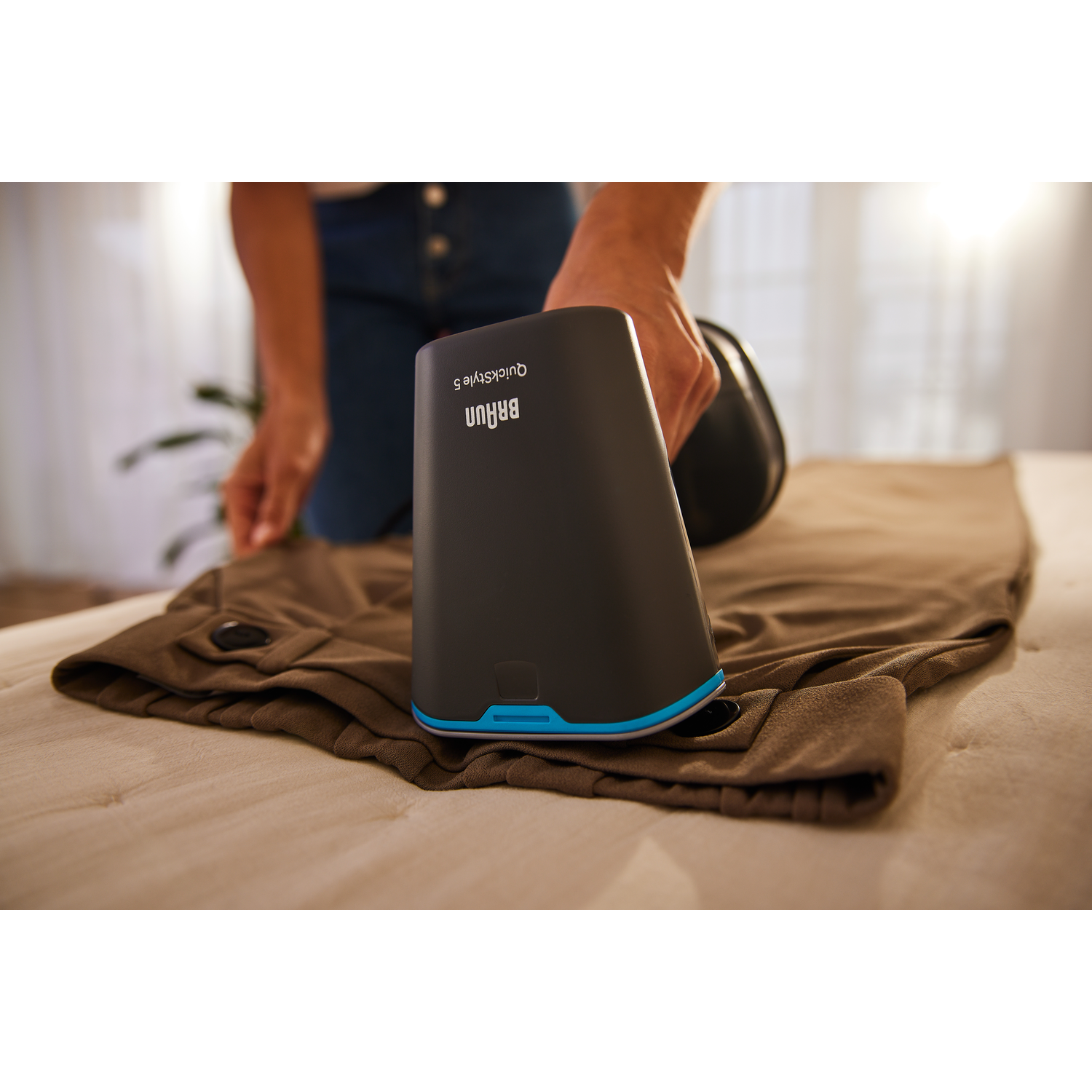Person using a Braun QuickStyle 5 on pants. Black device, blue accents, beige fabric.