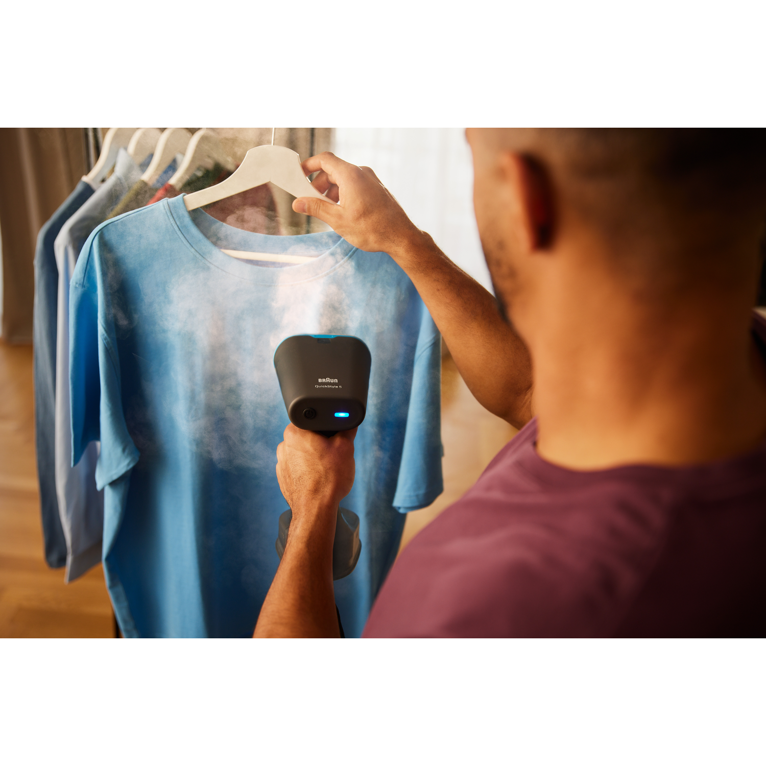 A man steaming a blue t-shirt with a garment steamer. The background has clothes on hangers.