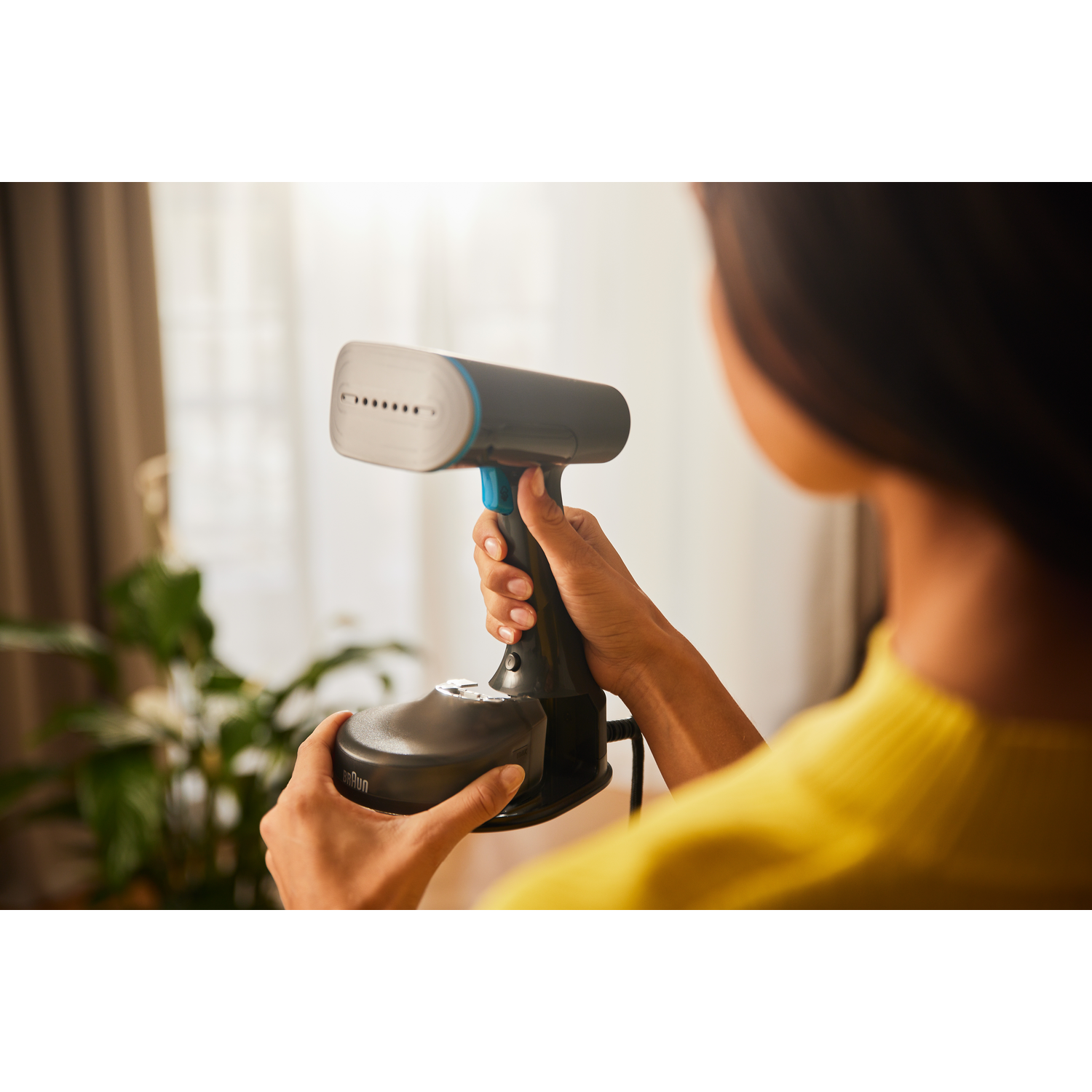 A person is using a hand steamer. The steamer is black and blue. A plant is in the background.