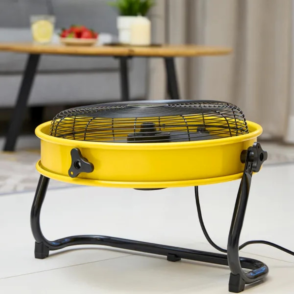 Żółty wentylator przemysłowy z czarną klatką, na białej podłodze. Tło salonu.