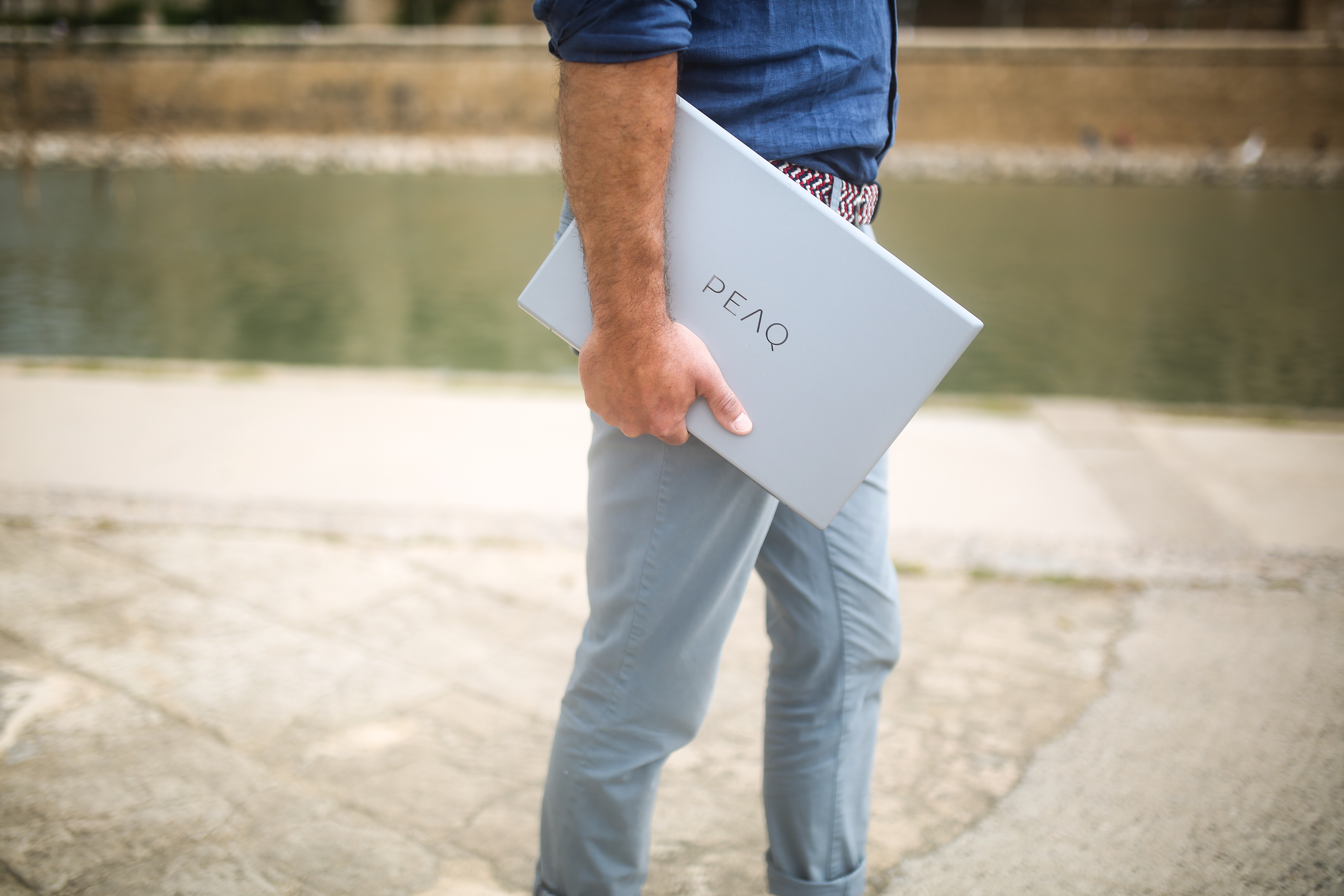 Ein Mann in blauem Hemd und grauer Hose hält einen grauen Laptop mit dem Wort PEAQ.