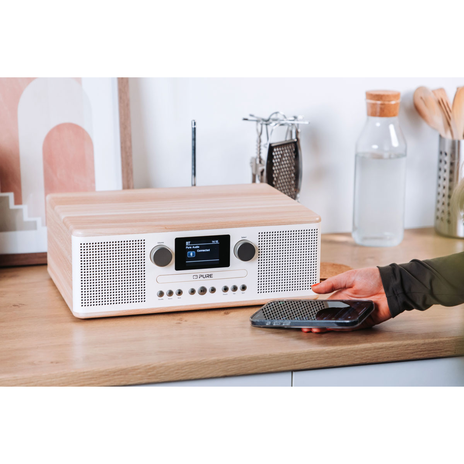 Een Pure-radio op een houten aanrecht met telefoon en accessoires, hand houdt de telefoon vast.
