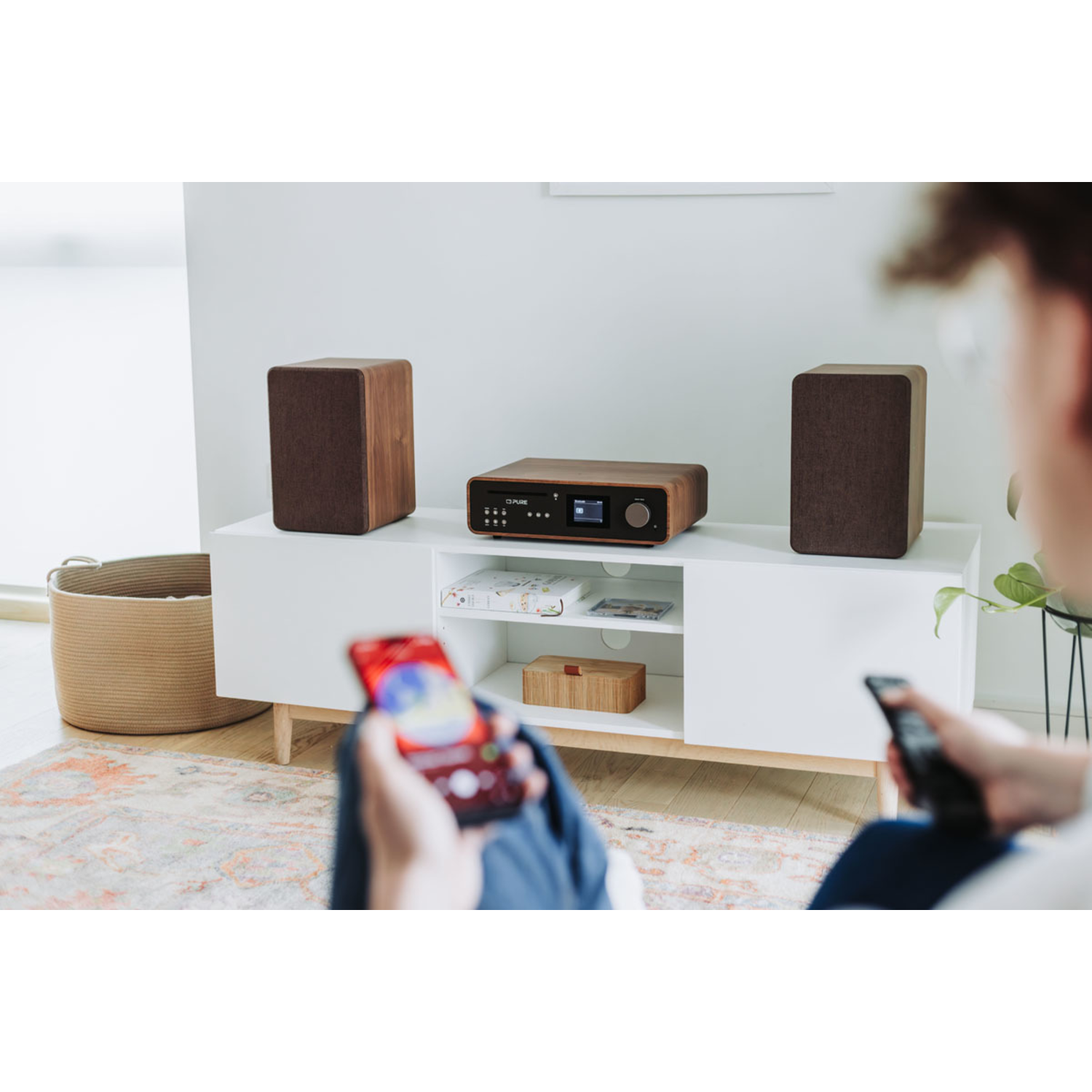 A person holding a phone and a remote, near a stereo system on a white cabinet.