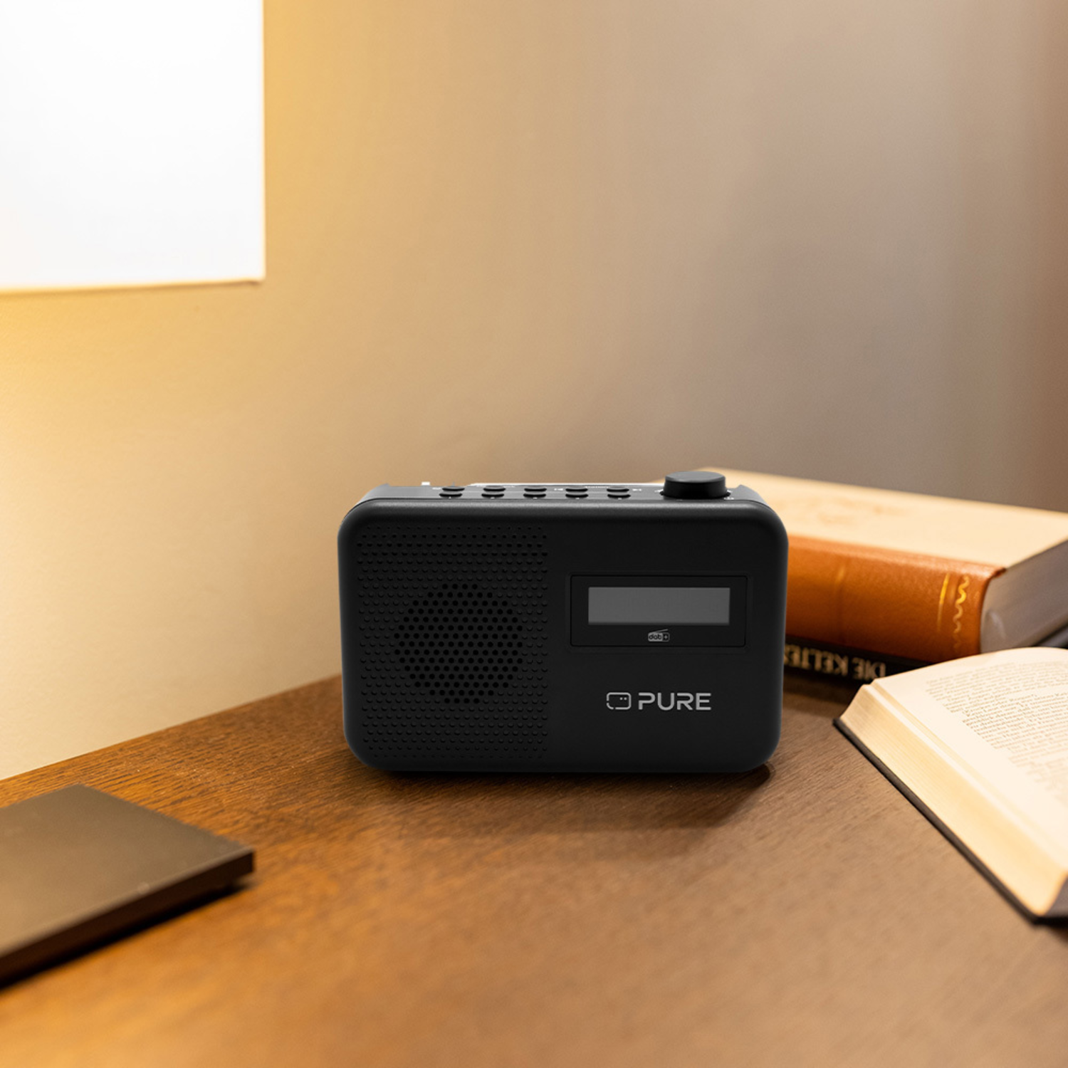 Black radio on a wooden table, near an open book and a lamp.