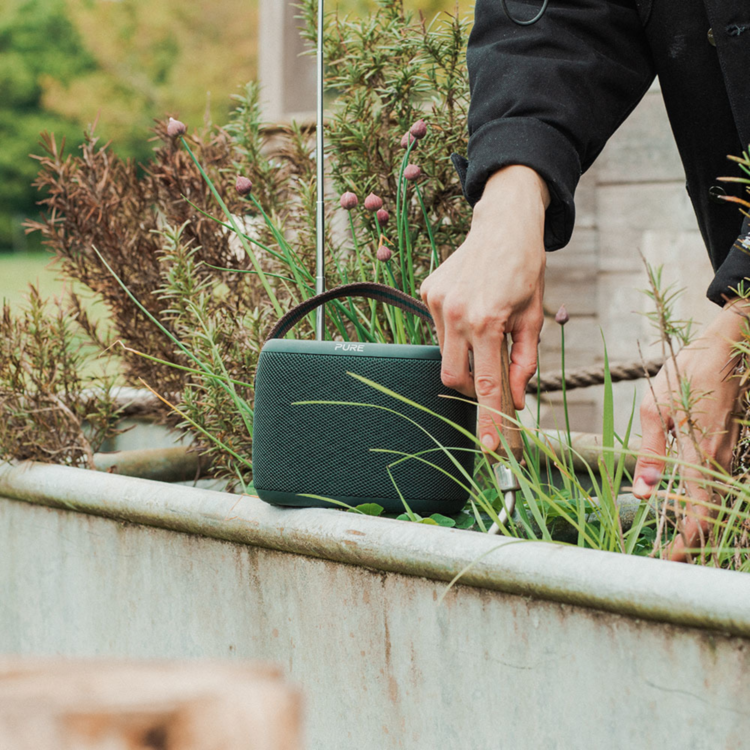 De hand van een persoon plaatst een groene draagbare luidspreker in een plantenbak met groen en bloemen.