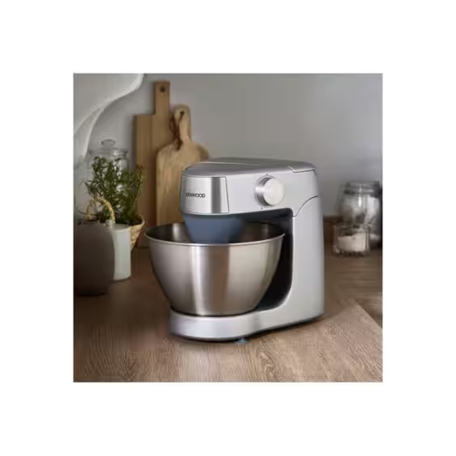 Een zilveren Kenwood keukenmixer op een houten aanrecht. Houten snijplanken op de achtergrond.