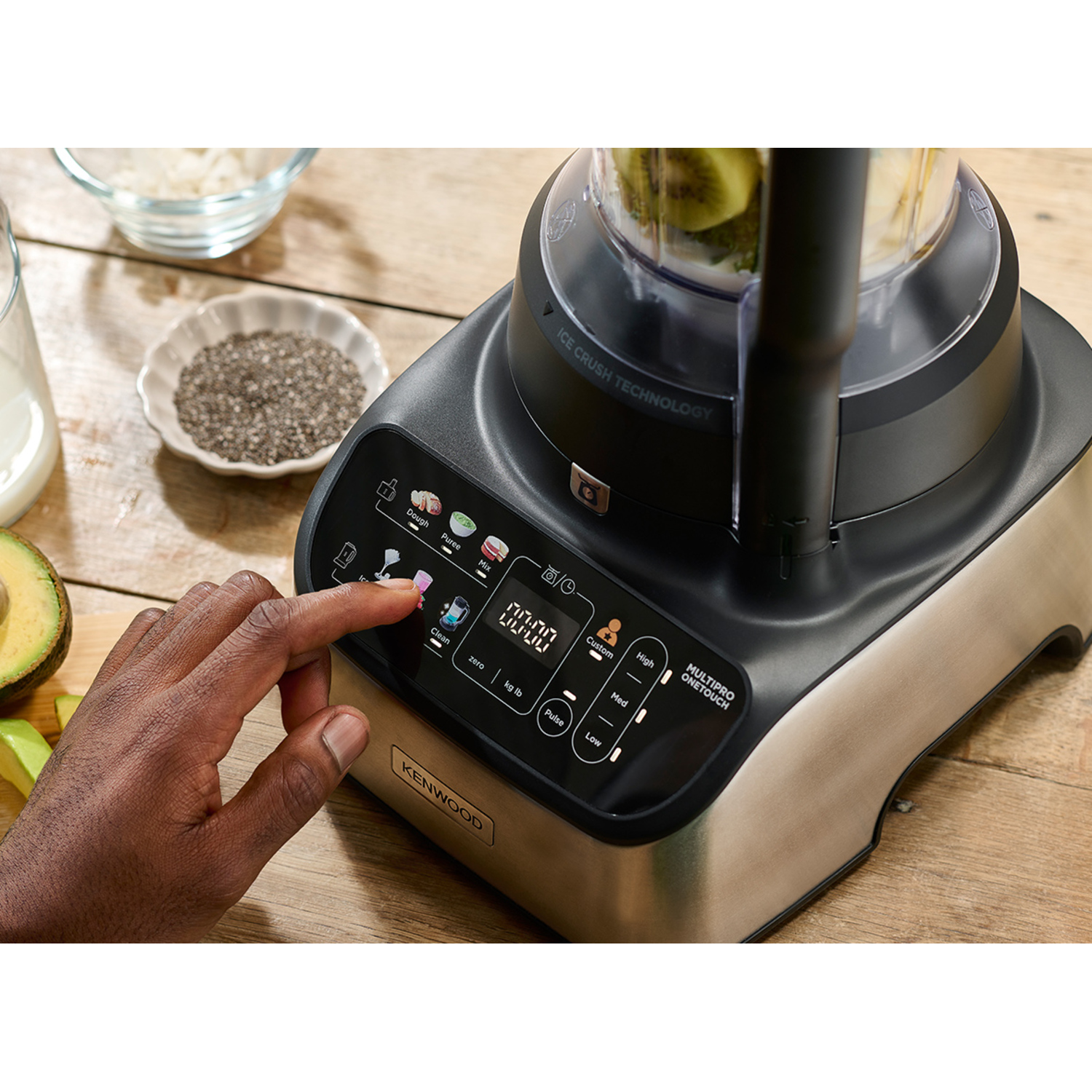 A food processor is on a wooden table. A hand is pressing a button. Ingredients are nearby.
