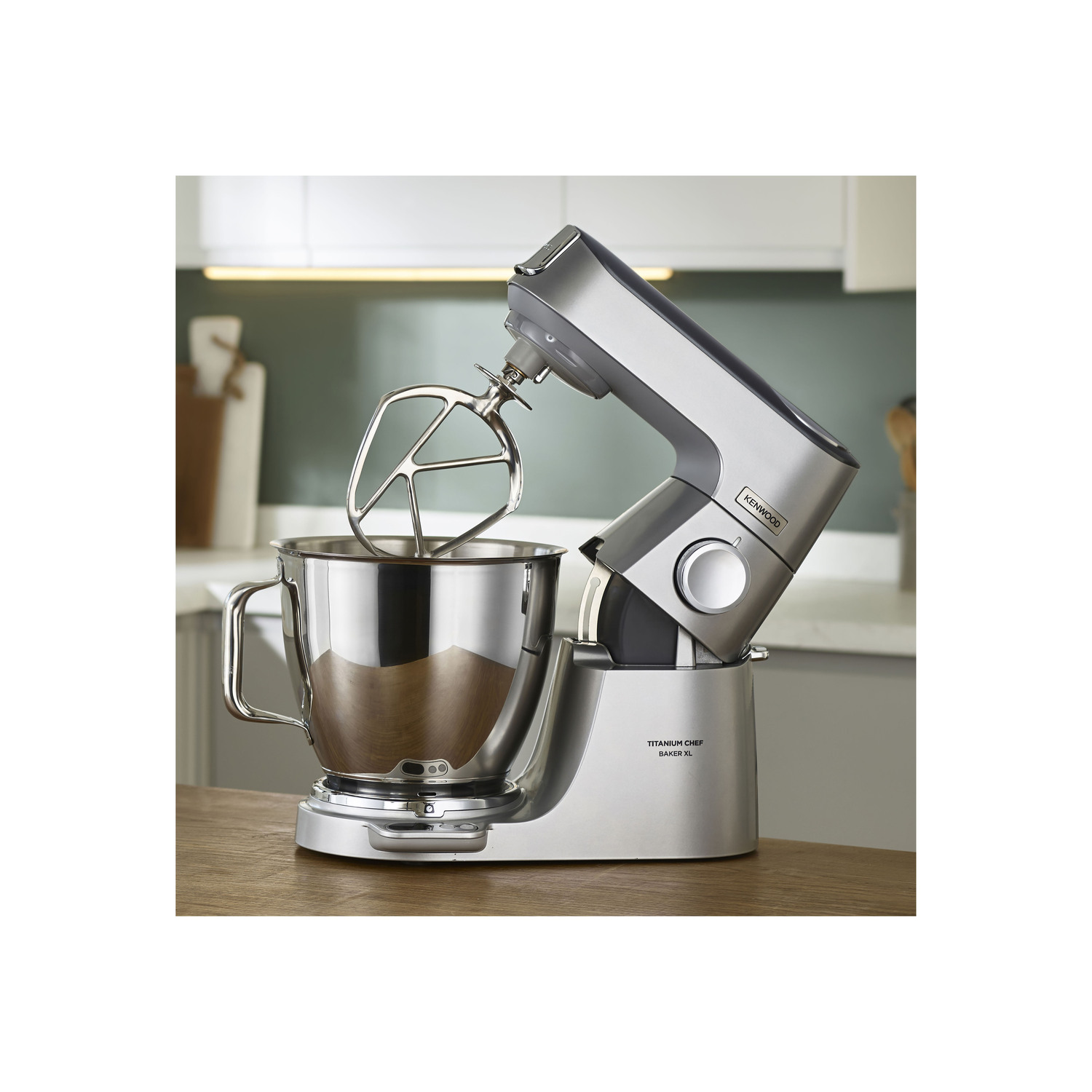Een zilveren en roestvrijstalen Kenwood Titanium Chef-mixer op een houten oppervlak. De kom en opzetstukken zijn zichtbaar.