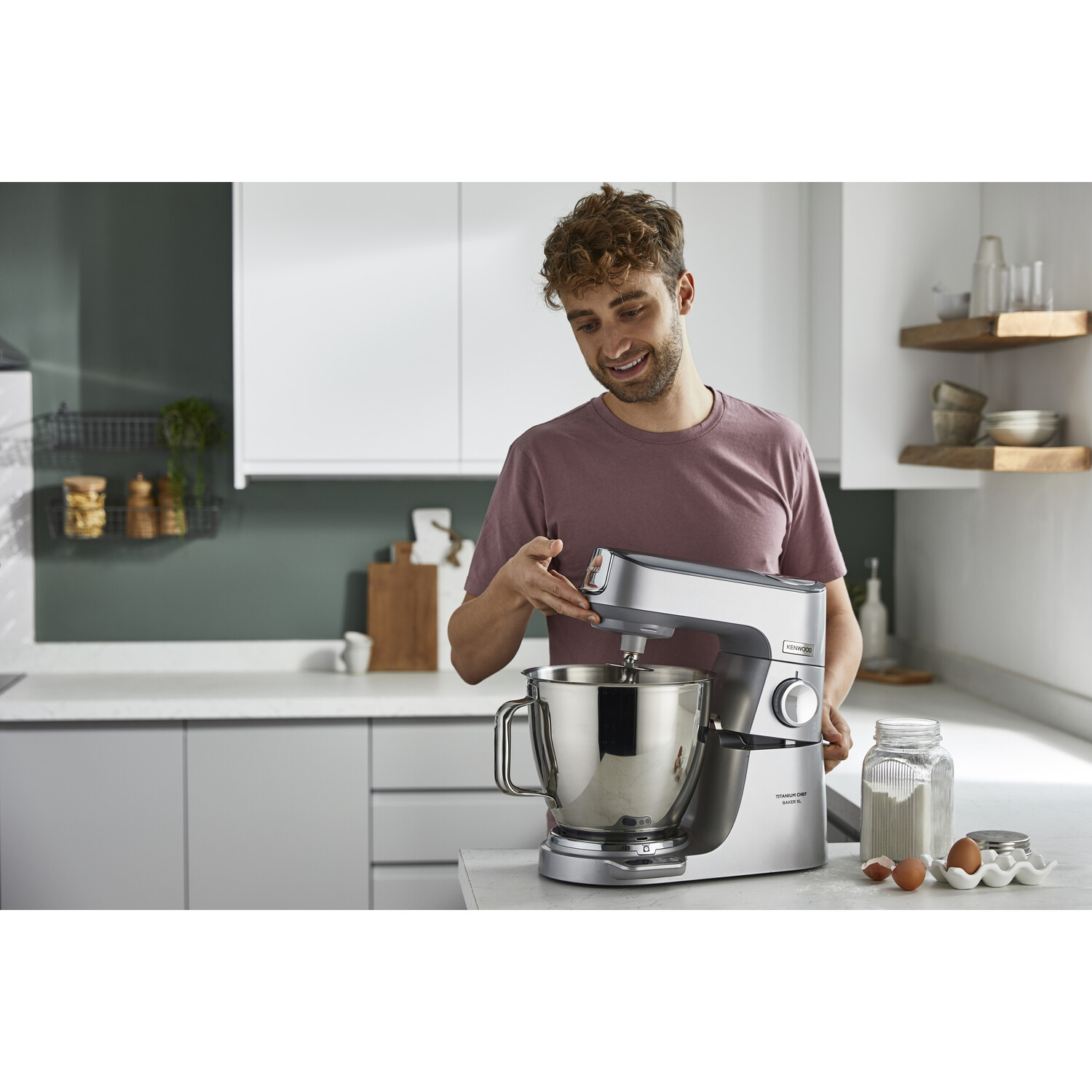 Een man gebruikt een zilveren keukenmixer in een moderne keuken, glimlachend. Ingrediënten zoals bloem en eieren liggen in de buurt.