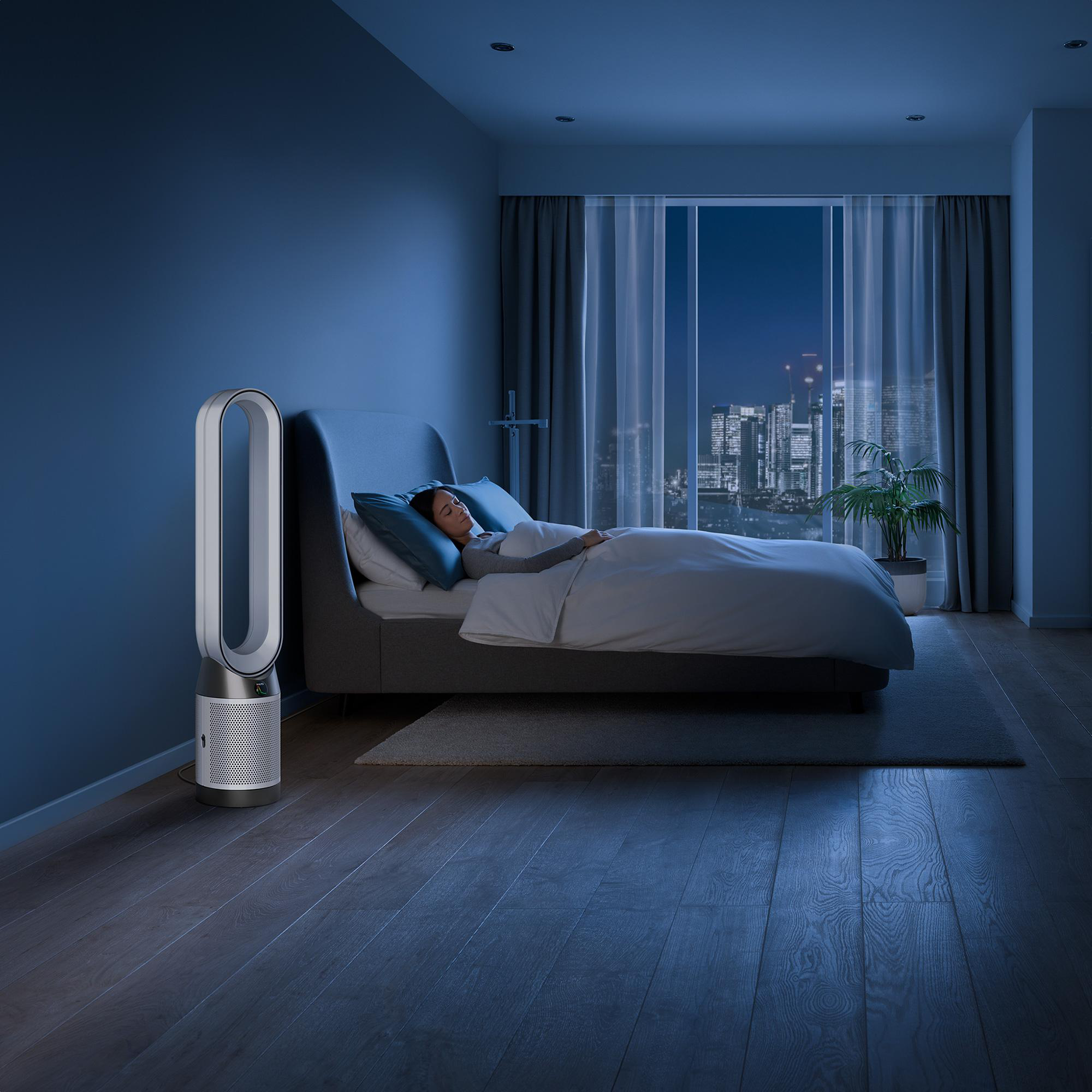 Eine Frau schläft in einem Bett, ein weißer Ventilator neben dem Bett, Nachtszene mit Stadtblick.