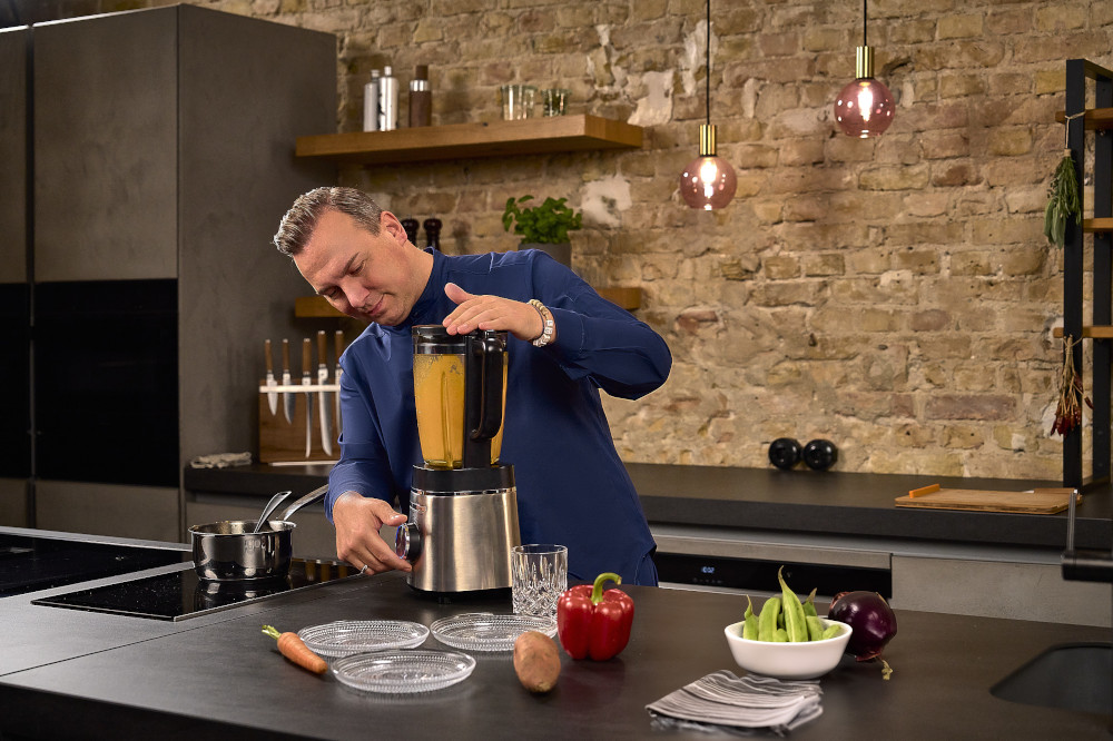 A man in a blue shirt blends a soup. Kitchen with brick wall and ingredients on the counter.