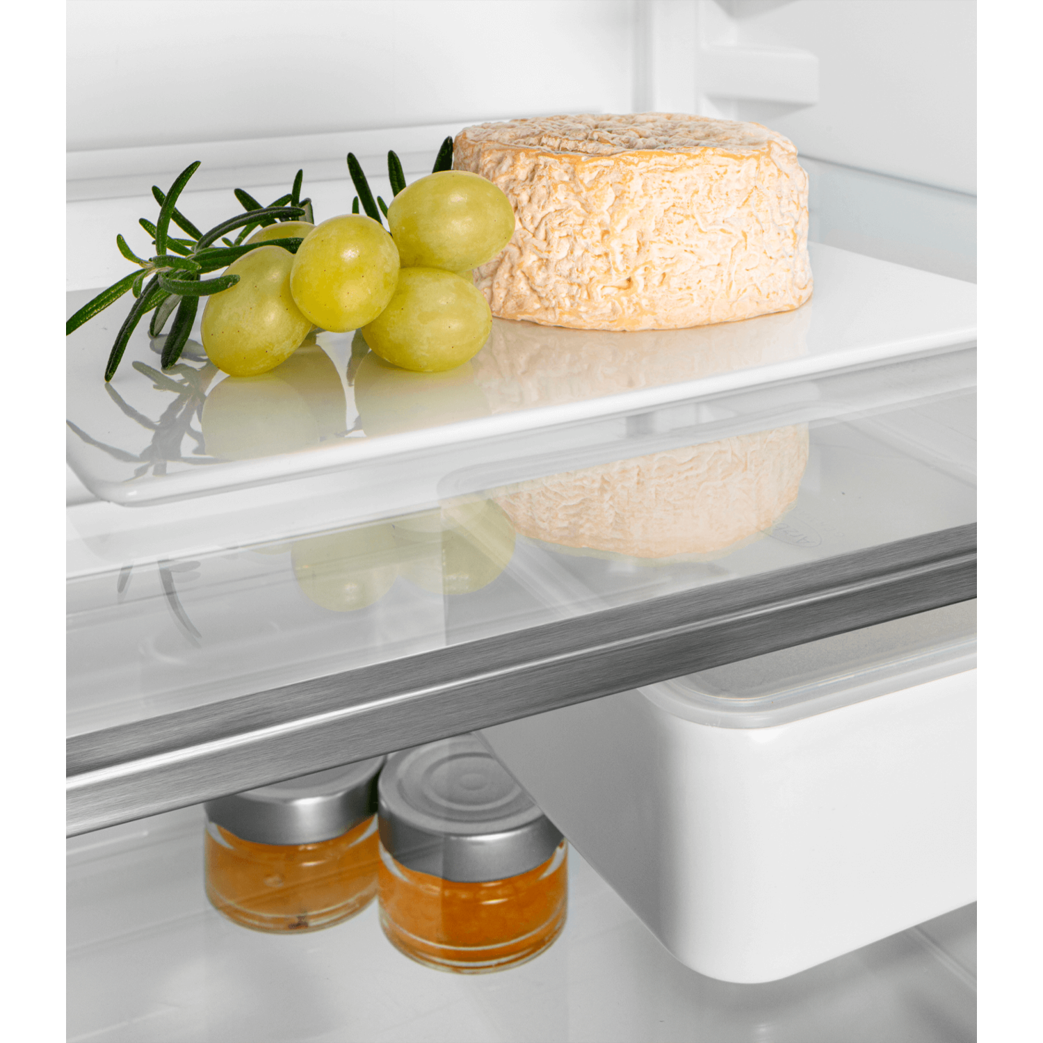 A fridge shelf with cheese, grapes, and rosemary. Jars of a spread are below.
