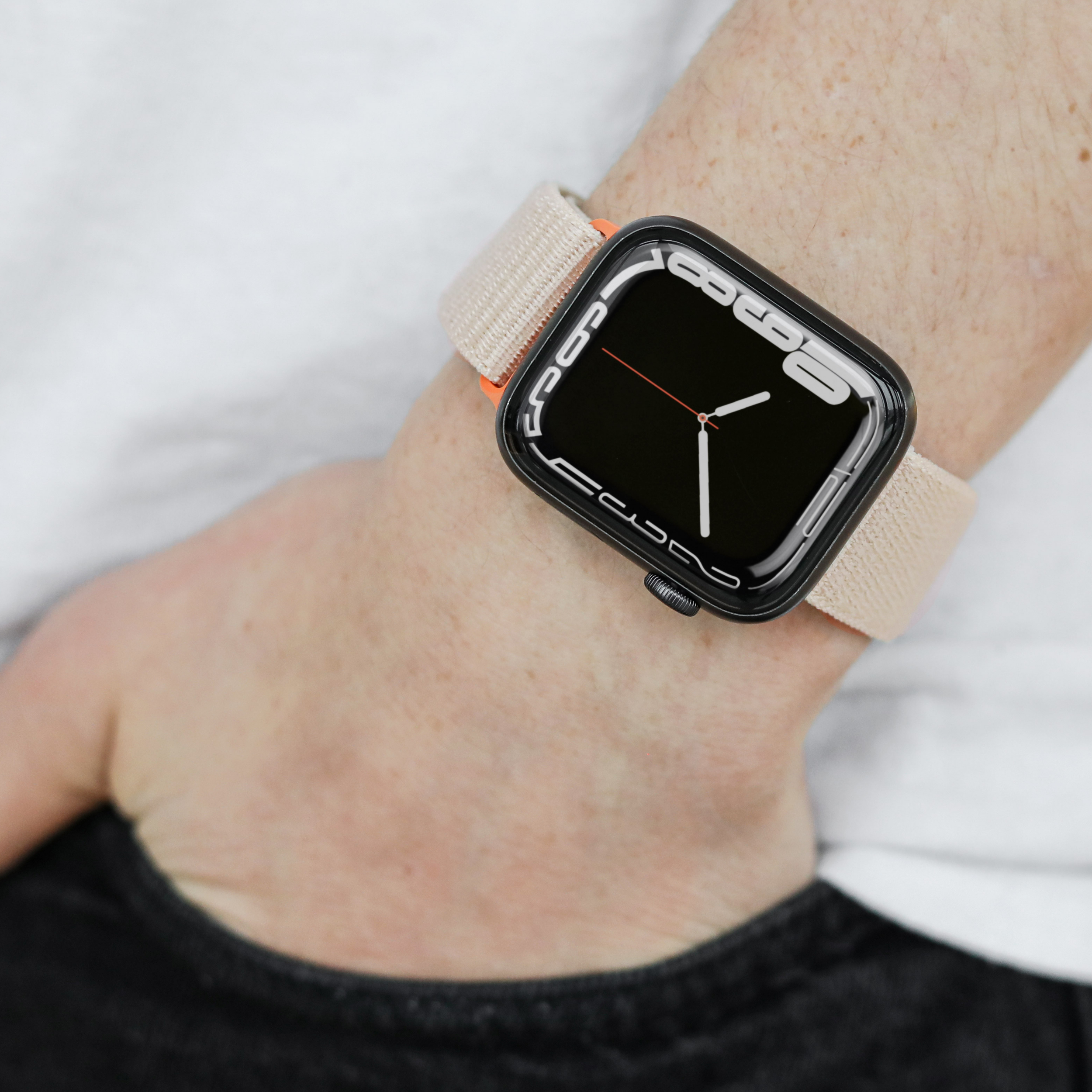 Das Handgelenk einer Person mit einer schwarzen Smartwatch. Die Uhr hat ein beiges Armband mit orangefarbenem Akzent.