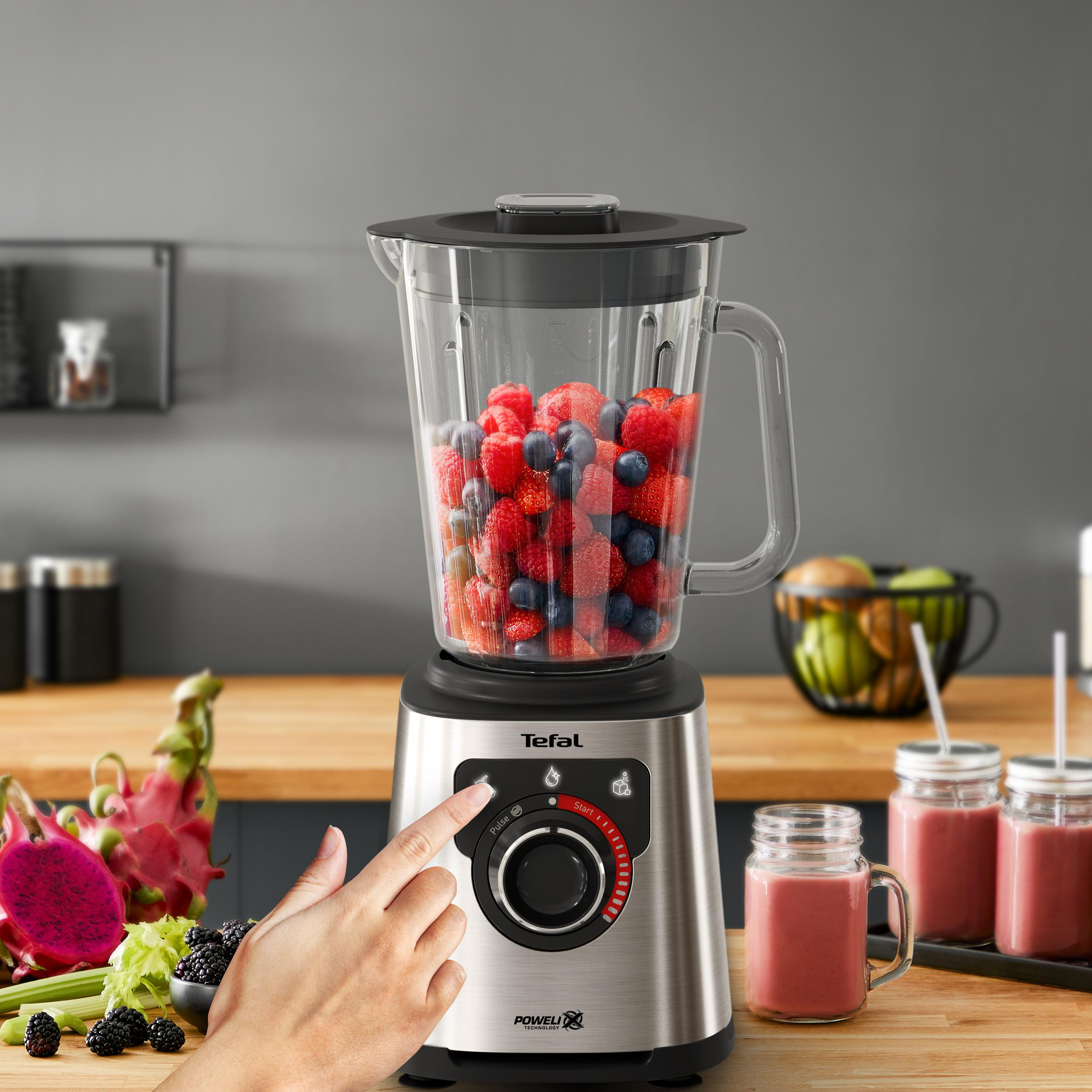 Mixer mit Beeren. Eine Hand drückt einen Knopf. Smoothie-Gläser daneben.