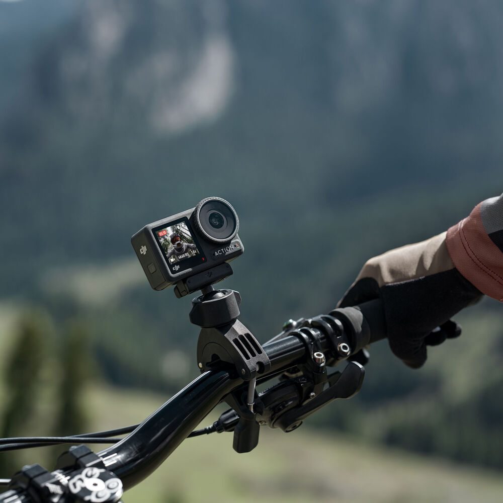 Action camera mounted on a bike handlebar. Person's hand on grip. Mountain scene in background.