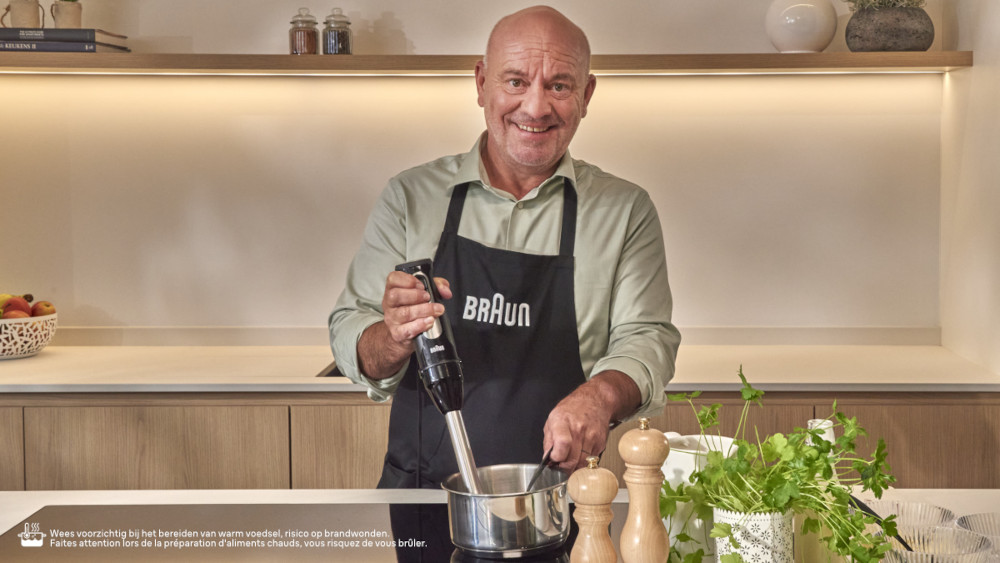 A man in a kitchen, holding a hand blender over a pot, smiling at the camera.