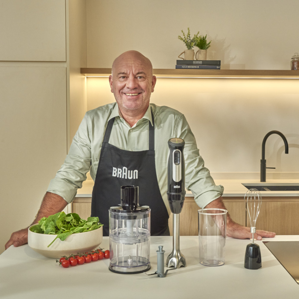 A man in a black apron stands in a kitchen, holding a blender and other kitchen tools.