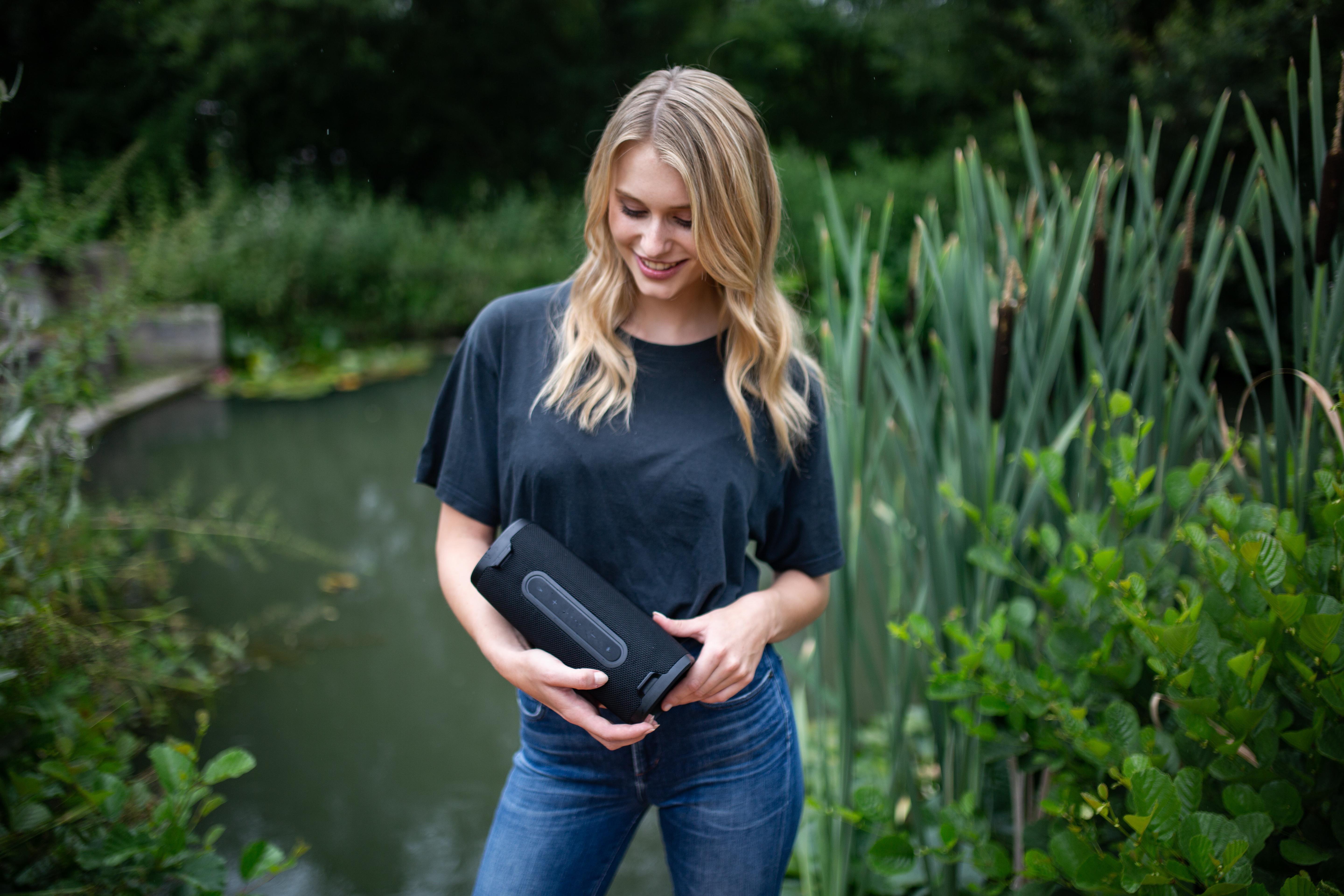 Frau hält einen schwarzen Bluetooth-Lautsprecher in einem Garten, mit Wasser.