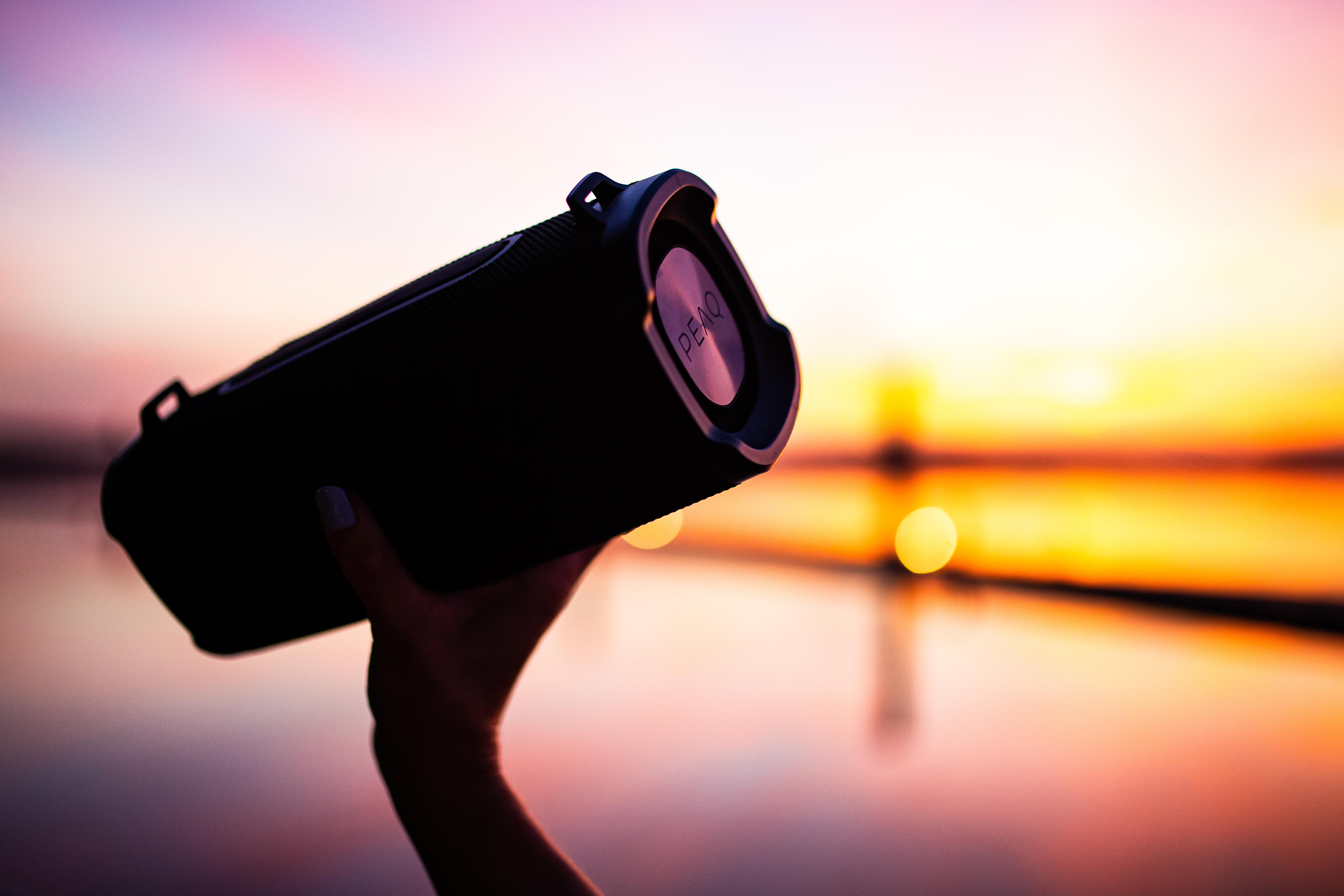 Hand hält einen schwarzen Lautsprecher mit silbernen Details. Sonnenuntergang im Hintergrund, der sich im Wasser spiegelt.