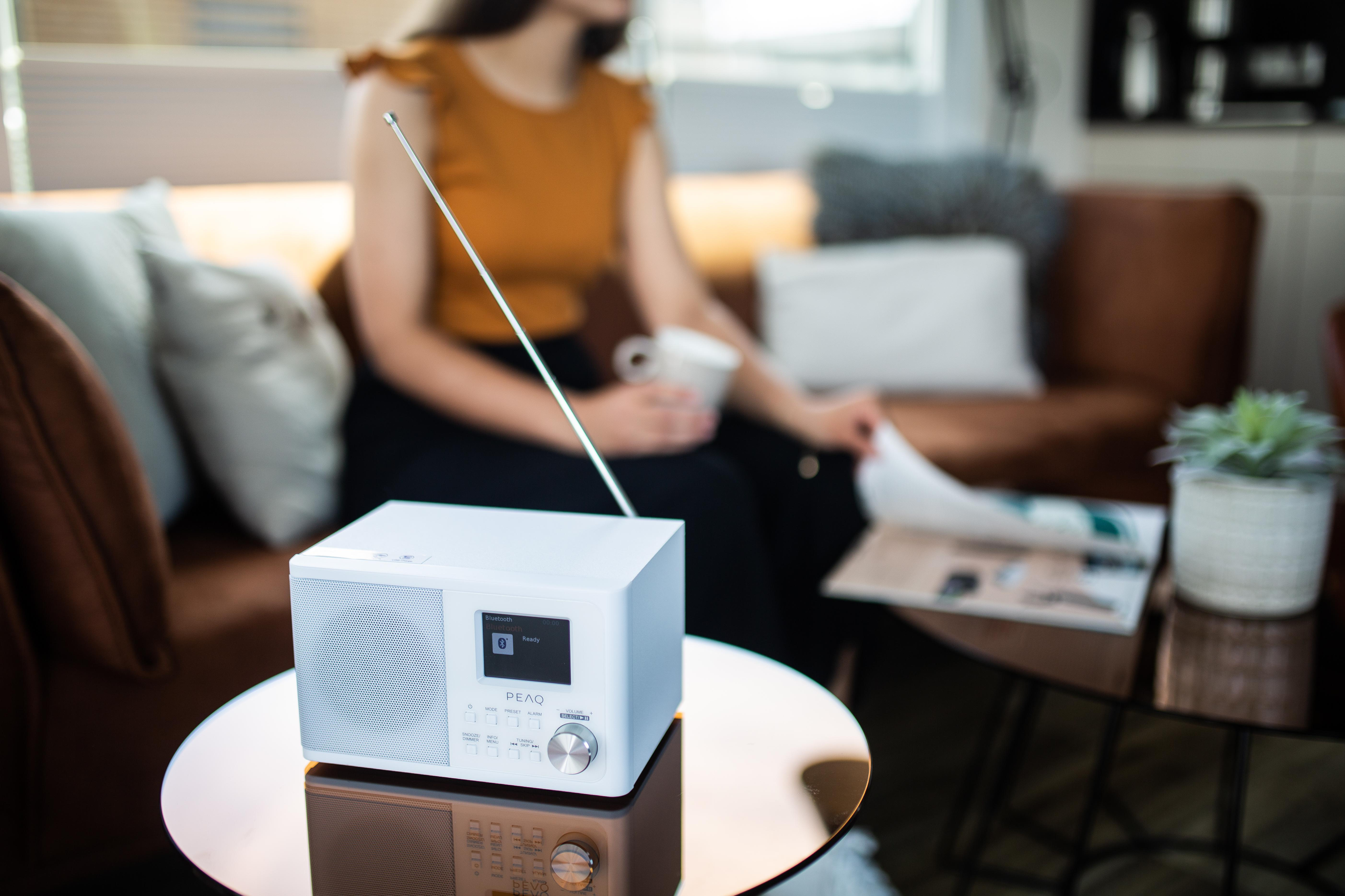 Ein weißes Radio steht auf einem Tisch. Eine Person sitzt dahinter und liest eine Zeitschrift.