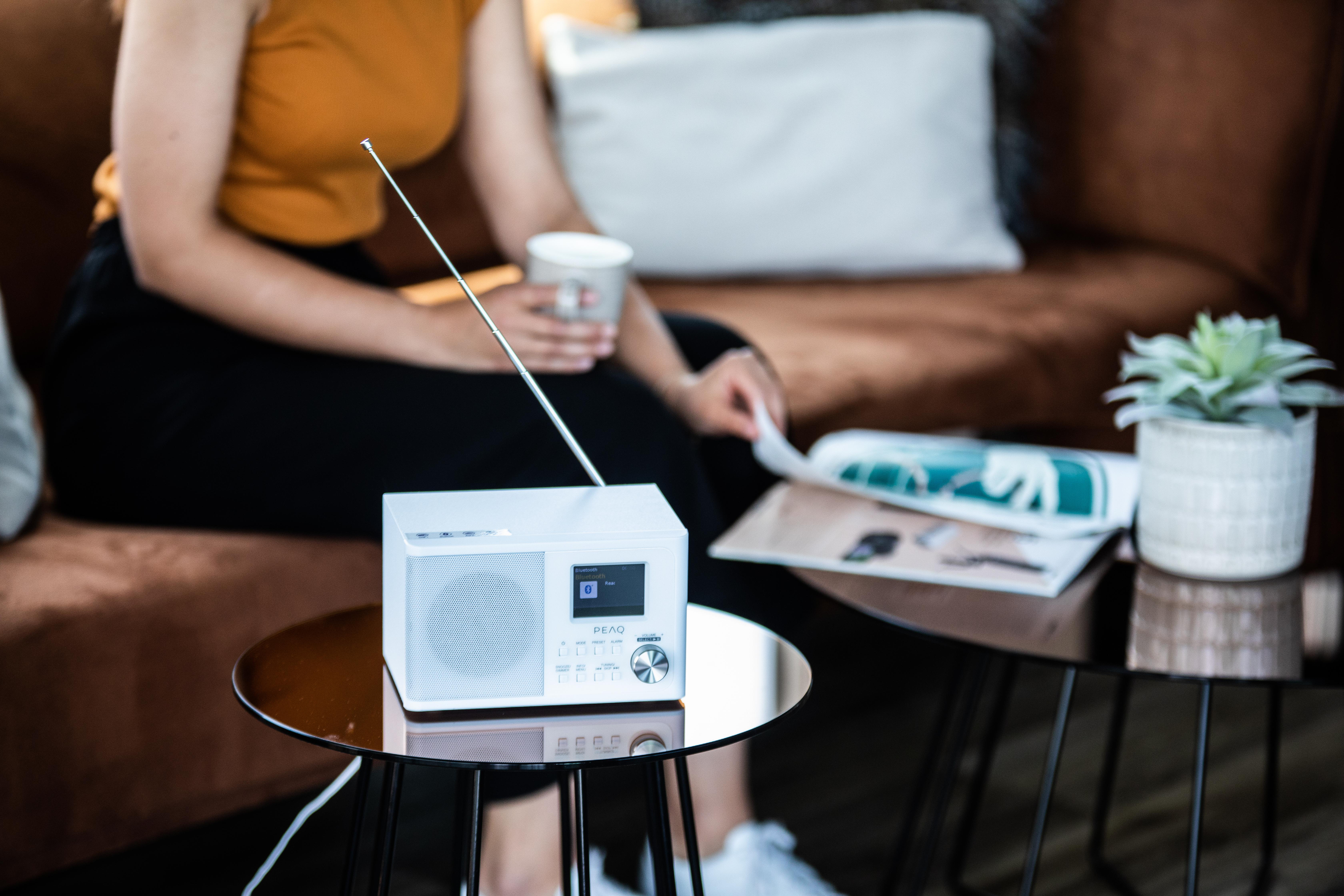 Ein weißes Radio auf einem Tisch. Eine Frau sitzt im Hintergrund auf einem Sofa und hält eine Zeitschrift.