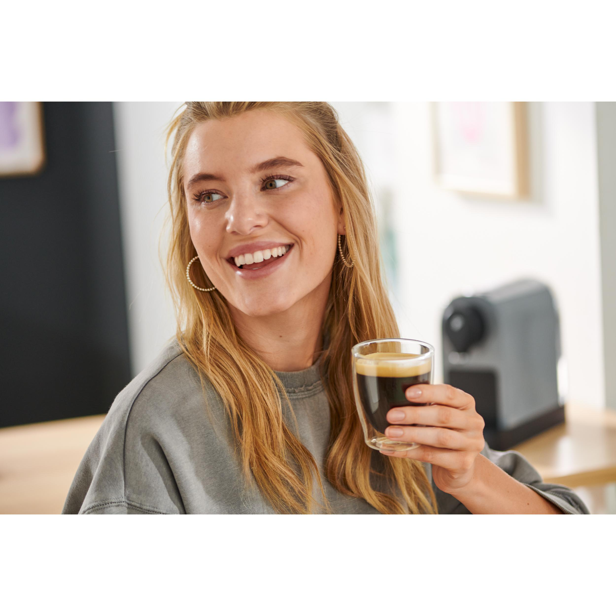 Eine lächelnde Frau, die ein Glas Kaffee in der Nähe einer grauen Kaffeemaschine hält.