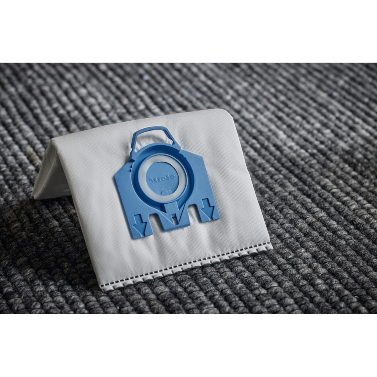 White vacuum cleaner bag with a blue Miele connector. Grey carpet background.