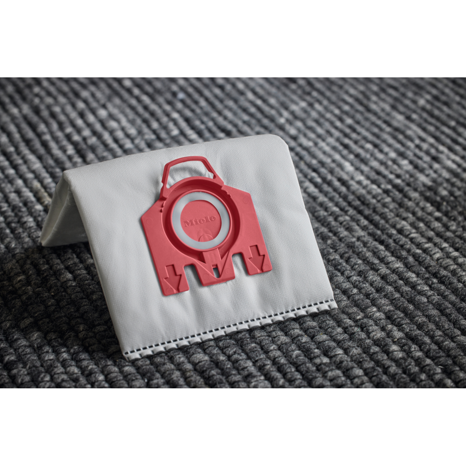 Gray vacuum cleaner bag with red Miele connector. The bag lies on gray patterned carpet.
