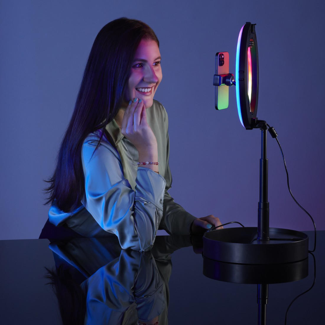 Eine Frau lächelt, während sie mit einem Telefon filmt, das an einem Ringlicht befestigt ist. Blaues und pinkes Licht.