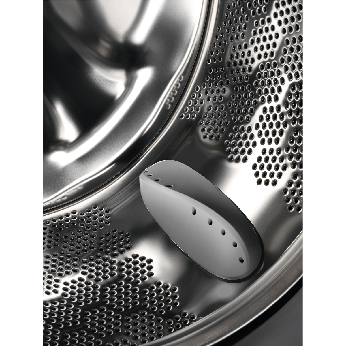 Inside a washing machine drum with holes, a white detergent dispenser is present.