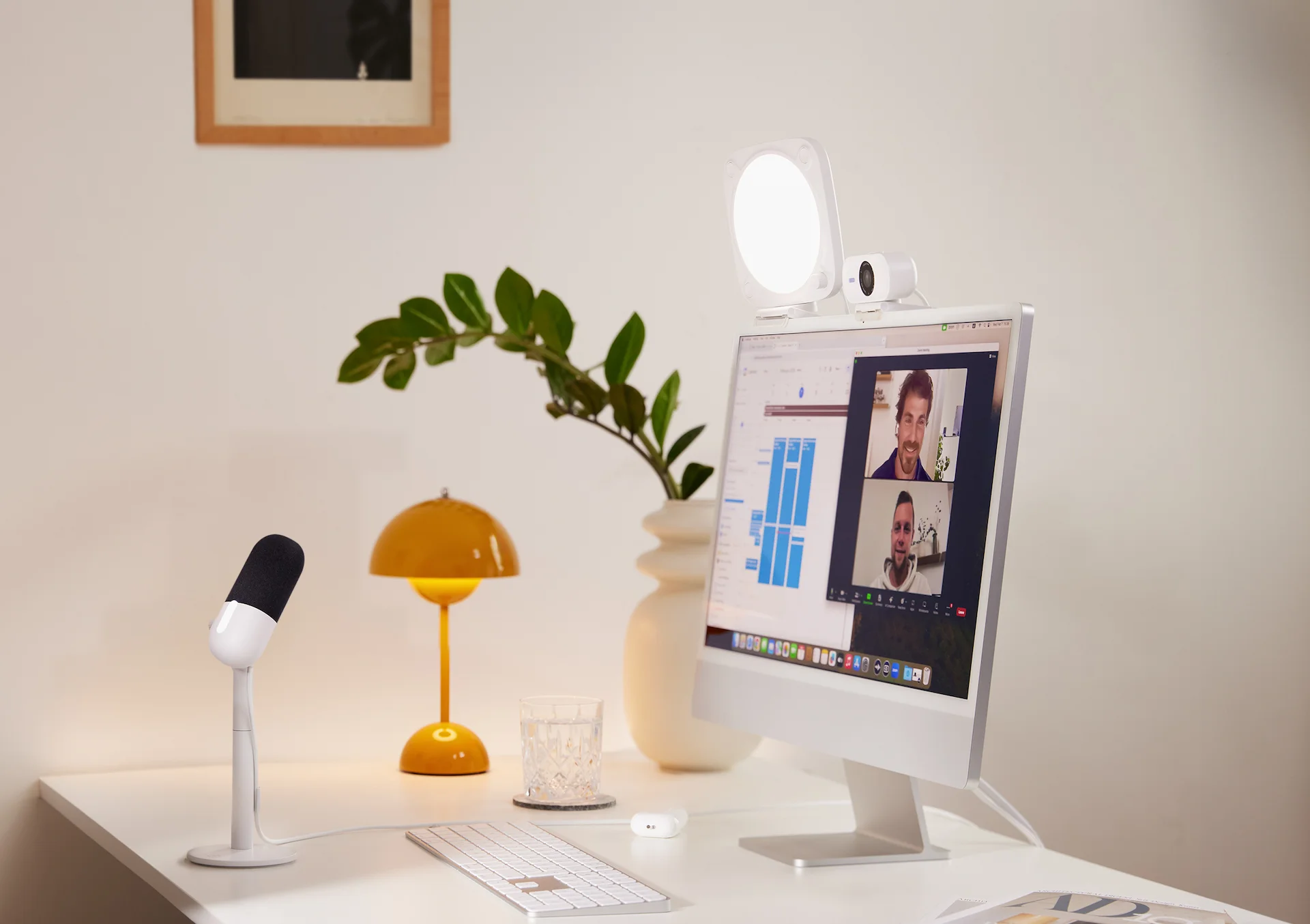 Schreibtisch mit Computer, der einen Videoanruf zeigt, Lampe, Mikrofon und Dekoration.